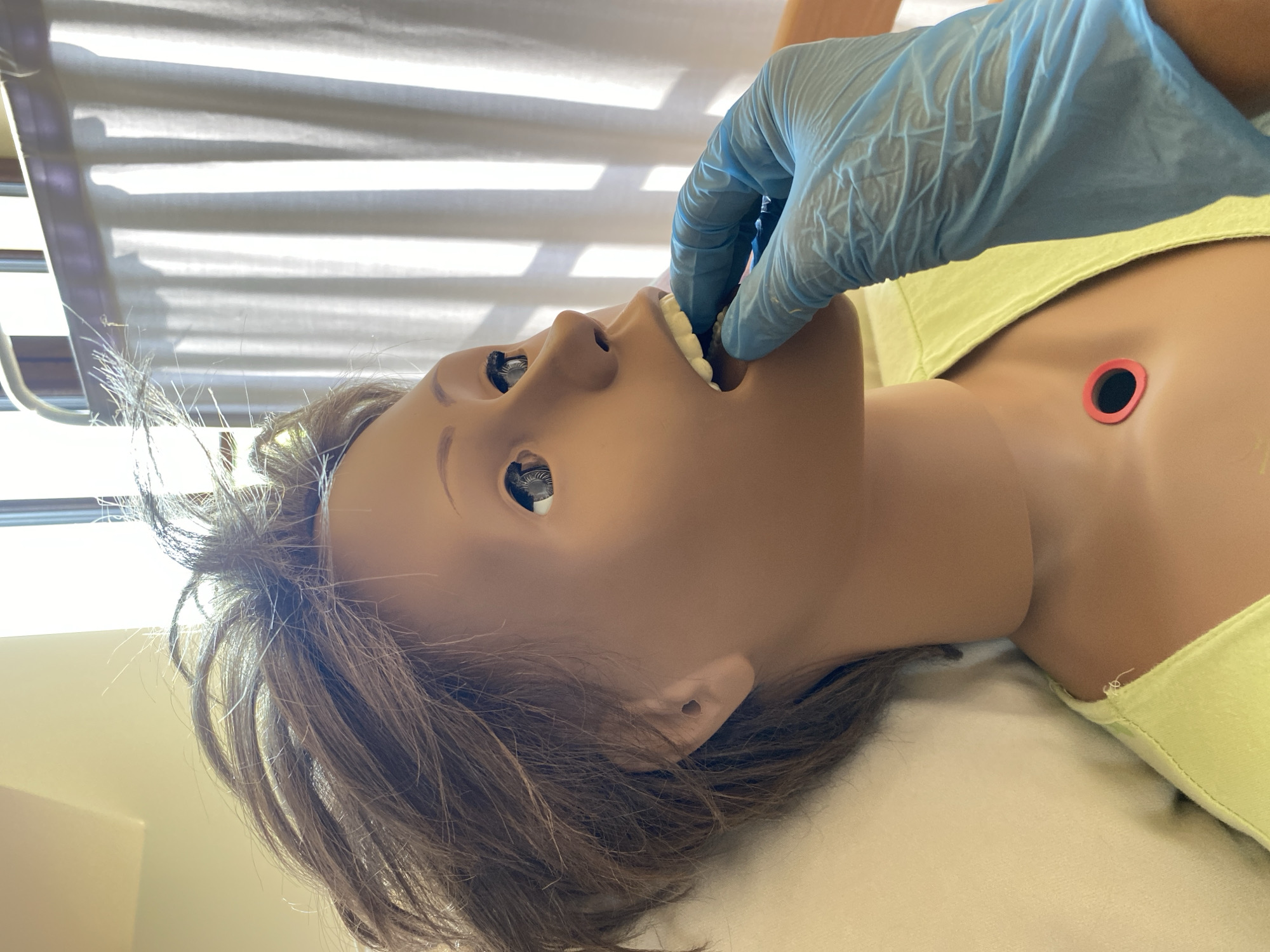 A close-up, eye-level shot shows a medical training mannequin's head and upper torso. The mannequin has brown hair, dark eyes, and a light brown complexion. A gloved hand, wearing a blue nitrile glove, is inserted into the mannequin's open mouth, pressing on its teeth. The mannequin is wearing a light green shirt, and a red and black tracheostomy tube is visible on its neck. The background consists of a white curtain and a window, suggesting an indoor setting, possibly a hospital or training facility. There is no indication of the city of Buk, Poland.