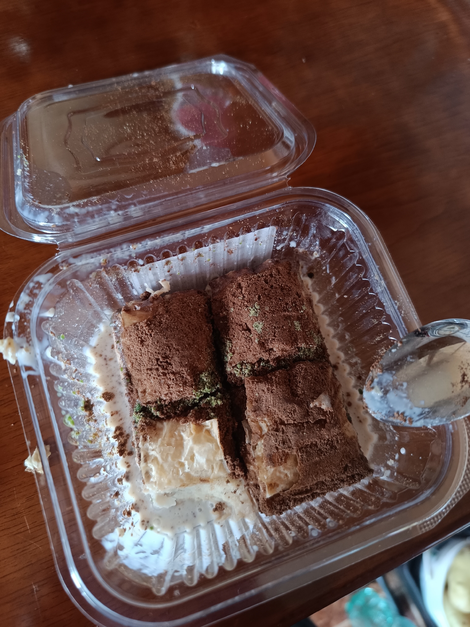 The image shows a partially eaten dessert in a clear plastic container with a detached lid. The dessert appears to be two rectangular pieces layered with a light-colored filling and heavily dusted with brown powder, possibly cocoa. Green specks are visible on top of the dessert. The container has traces of white liquid, likely cream, and crumbs. A spoon rests on the right side of the container with traces of the dessert. The container sits on a wooden surface. The location is unknown, but the user specified the city of Pleven, Bulgaria, which may or may not be relevant to the scene.