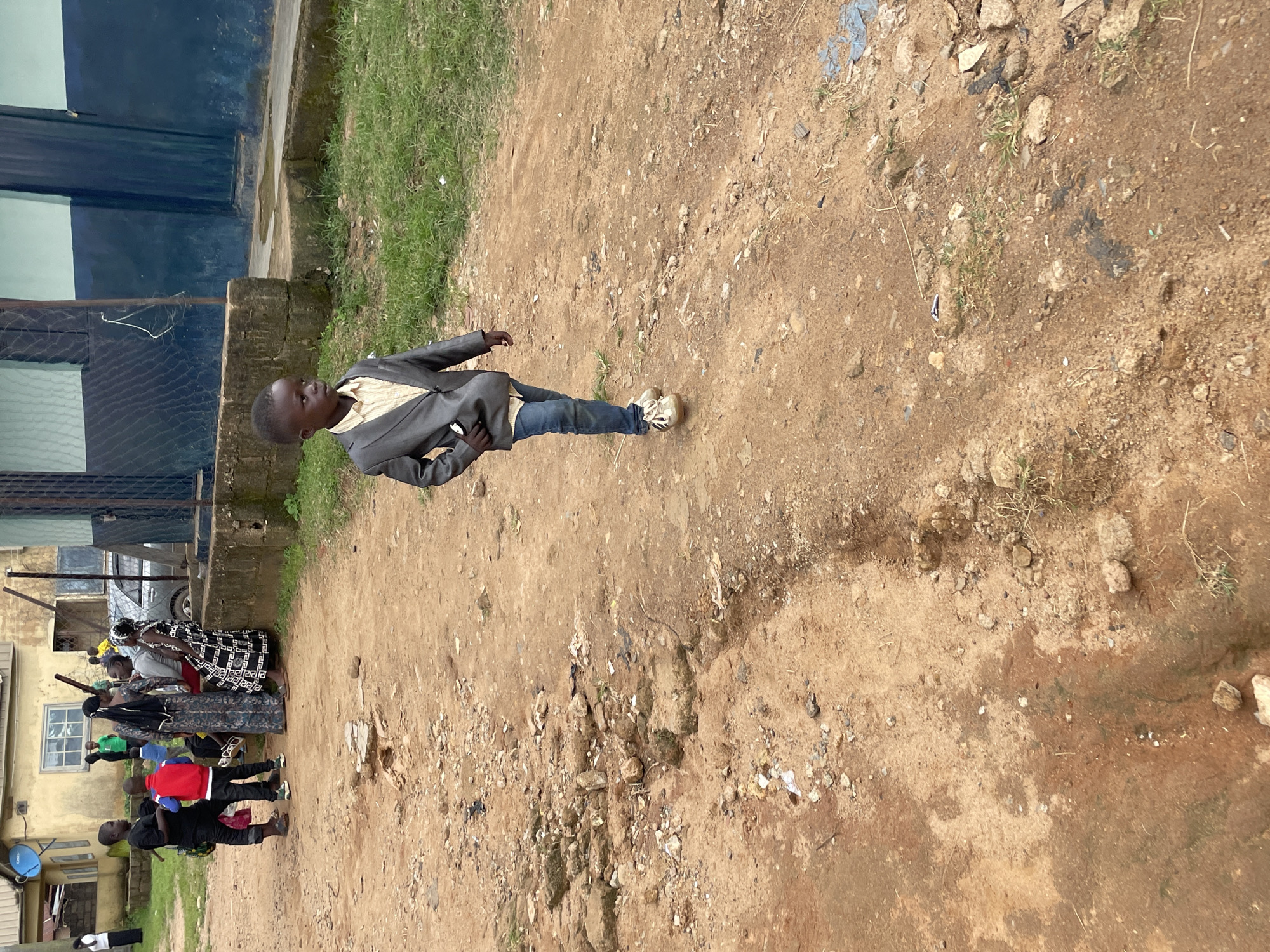 In Barakin Lerawi, Nigeria, a young boy, appearing to be of elementary school age, walks away from the viewer on a dusty, dirt road. He is wearing a light-colored collared shirt and a dark grey suit jacket, dark blue jeans, and tan sneakers. The road is uneven and littered with rocks and debris. In the background, several people are gathered near a building with a blue and light green facade. Some are children, one wearing a red jacket, and others appear to be adults dressed in casual clothing and traditional Nigerian attire. The environment suggests an outdoor urban setting, possibly a community area or a street in a residential part of the city.
