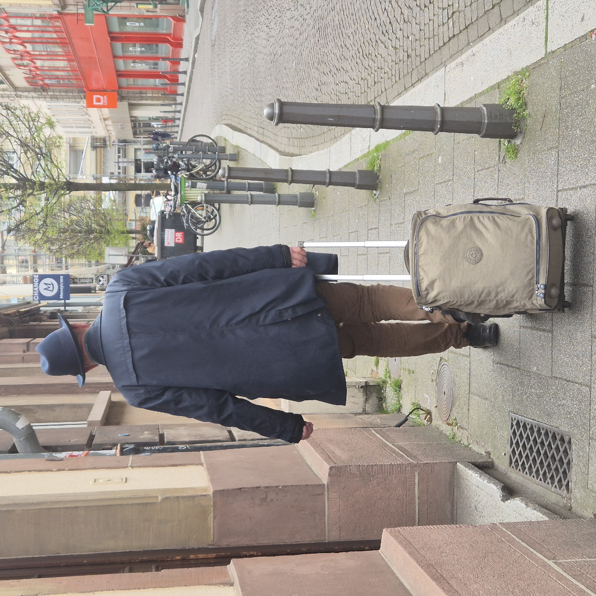 A man wearing a blue hat, a dark blue jacket, and brown pants is walking on a sidewalk in Strasbourg, France, pulling a beige rolling suitcase. He is walking past a building with reddish-brown stone steps and a drain. Metal bollards separate the sidewalk from a cobblestone street where several bicycles are parked. Further down the street, there is a red building with the letter "D" in a square on its facade and a sign for a money exchange. A tree with sparse green leaves is also visible.