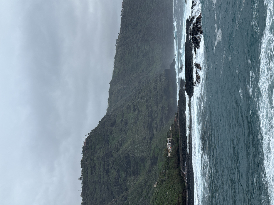 A mountain covered in lush green vegetation meets a rocky coastline. Waves crash against the rocks, creating white foam on the surface of the dark blue water. A small building stands on the edge of the mountain. The sky is overcast with gray clouds. The photograph is slightly tilted, with the mountain running vertically on the left.