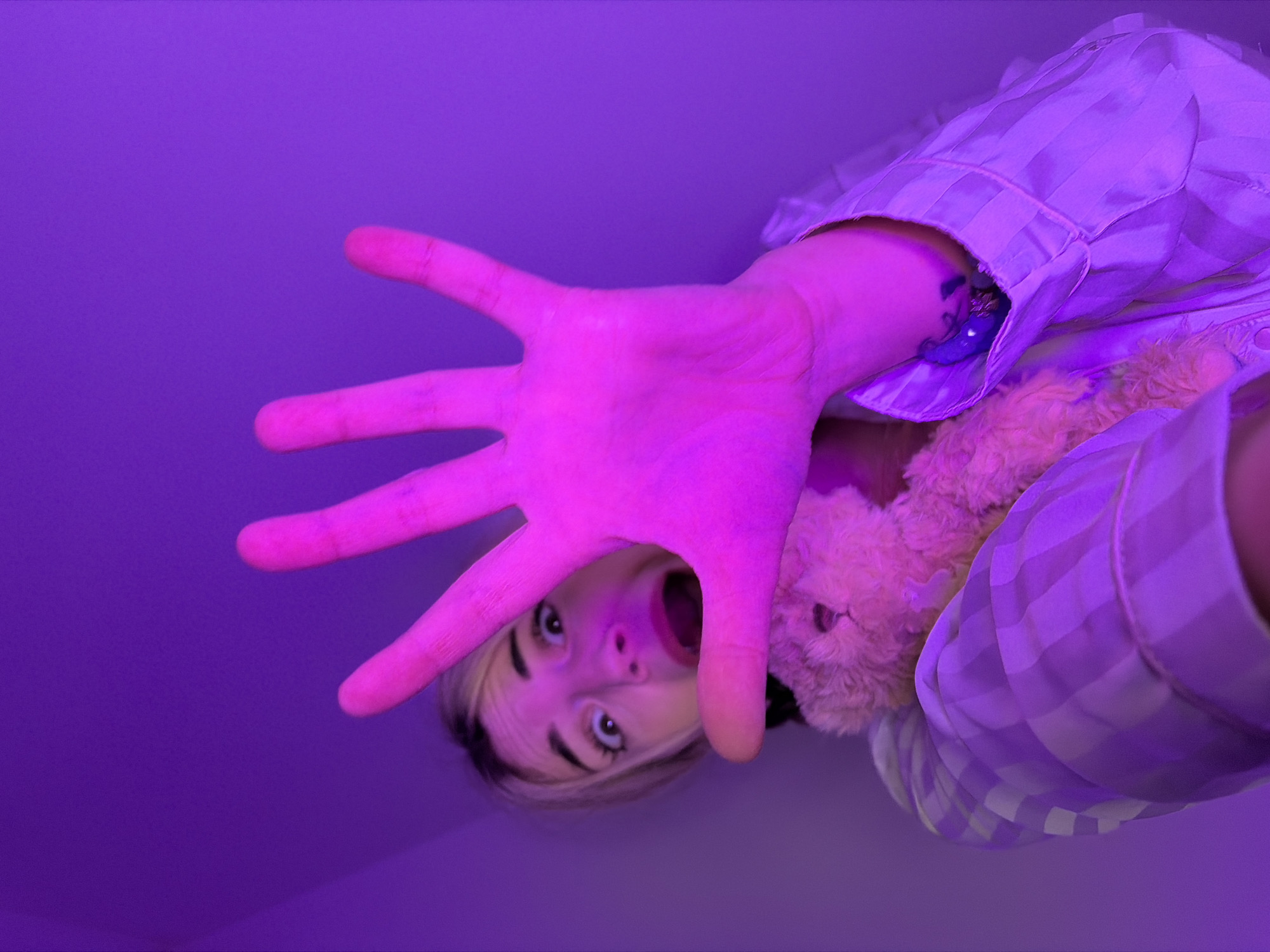 The image depicts a close-up, low-angle, indoor scene illuminated by intense purple ambient light.
**Location:** No specific location within Budva, Montenegro, or any other city can be determined from the visual evidence. The background appears to be an interior wall or ceiling, uniformly colored purple by the lighting.
**People:** One female individual is present, positioned in the lower portion of the frame, looking upward towards the camera. She has light-colored hair, possibly blonde, with darker roots visible around her face. Her eyes are wide open, and her mouth is agape in an expression consistent with surprise or alarm.
**Subjects/Objects:**
* **Hand:** The individual's right hand is fully extended forward, towards the camera, with the palm open and fingers splayed, occupying a significant portion of the central frame. The intense purple lighting renders the hand magenta.
* **Plush Toy:** A small, light-brown or beige plush toy, resembling a teddy bear, is visible resting on her left shoulder/chest area, partially obscured by her extended hand.
* **Clothing:** She is wearing a light-colored, long-sleeved shirt or pajama top with a faint vertical striped pattern.
* **Left Arm/Wrist:** On her left wrist, a small, dark tattoo is visible just above the cuff of her shirt. Below the tattoo, a dark, possibly metallic or beaded object, such as a bracelet or charm, is partially visible.
**Actions/Interactions:** The individual is actively presenting her open hand towards the camera, creating a direct interaction with the viewer. Her facial expression suggests a startled or reactive state to something immediately in front of her or the camera's position.
**Environment:** The entire scene is bathed in a strong, artificial purple light, creating a monochromatic effect. The background is featureless and smooth, suggesting an interior wall or ceiling without discernible architectural details.