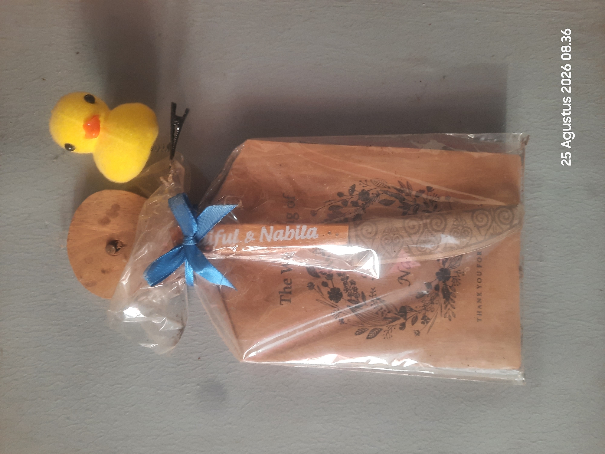 An overhead shot shows a wedding favor wrapped in clear plastic. The favor is a flat wooden item, possibly a spoon or spreader, decorated with intricate patterns and text that reads "Fulf & Nabilla" vertically along its length. Below this, the text "The Wedding of" and "Na" are visible, along with floral and swirling motifs on brown paper backing. A blue ribbon is tied in a bow at the top of the plastic wrap. To the right of the favor, a small yellow plush duck toy with black eyes and an orange beak is attached to the plastic wrap by a black hair clip. The favor and duck are hanging on a light grey textured wall, with a wooden disc visible behind the top of the favor. The bottom right corner of the image displays the date and time "25 Agustus 2026 08.36".