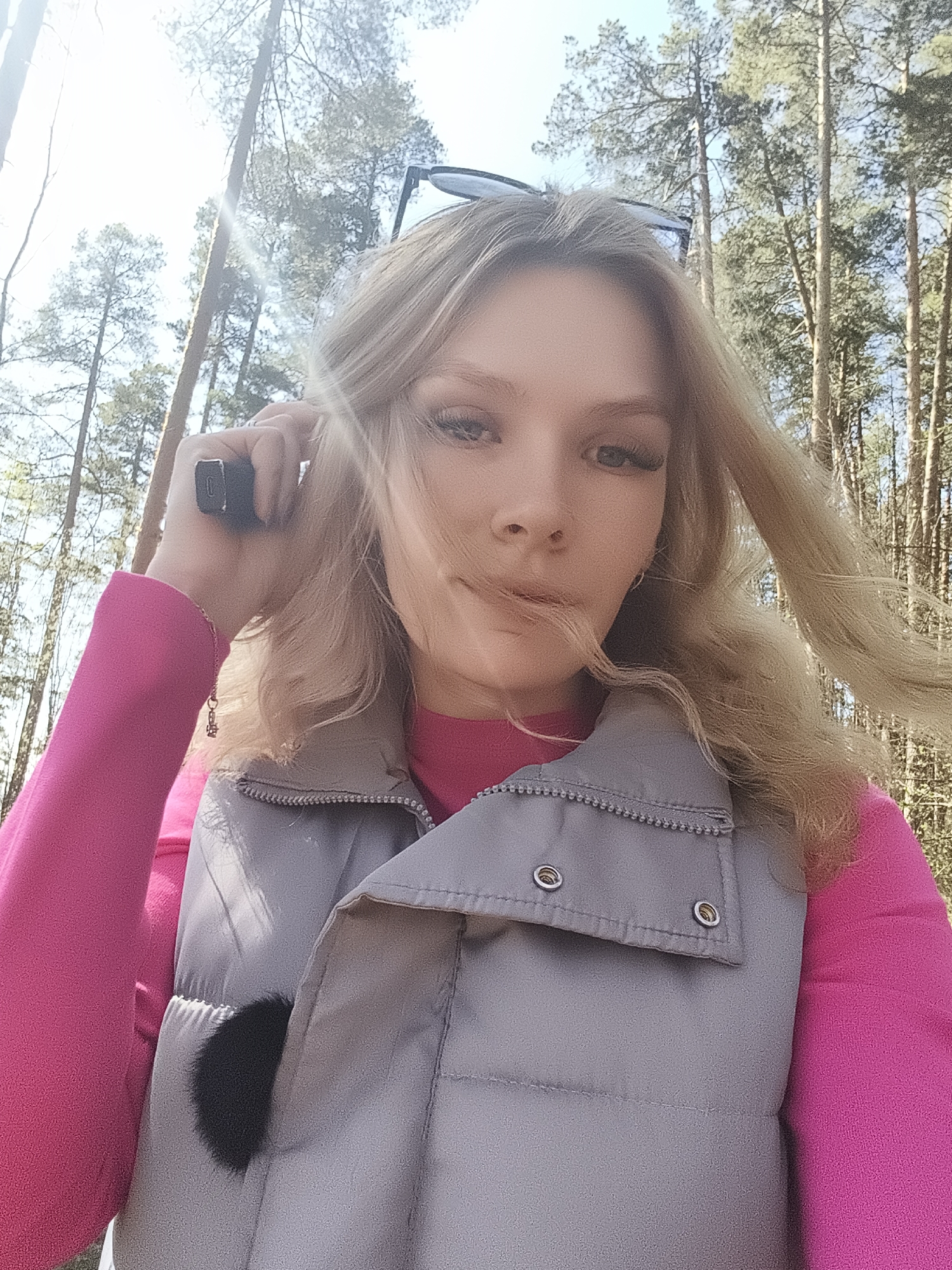 A fair-skinned female with blonde, shoulder-length wavy hair is shown in a selfie outdoors. She is wearing a pink long-sleeved shirt and a gray vest with a fur pom-pom visible in the pocket. Sunglasses are perched atop her head. She holds a rectangular object to her right ear. Tall, slender trees form a blurred background, suggesting a forest or wooded area. The specific location within Izhevsk, Russia cannot be determined from the image.
