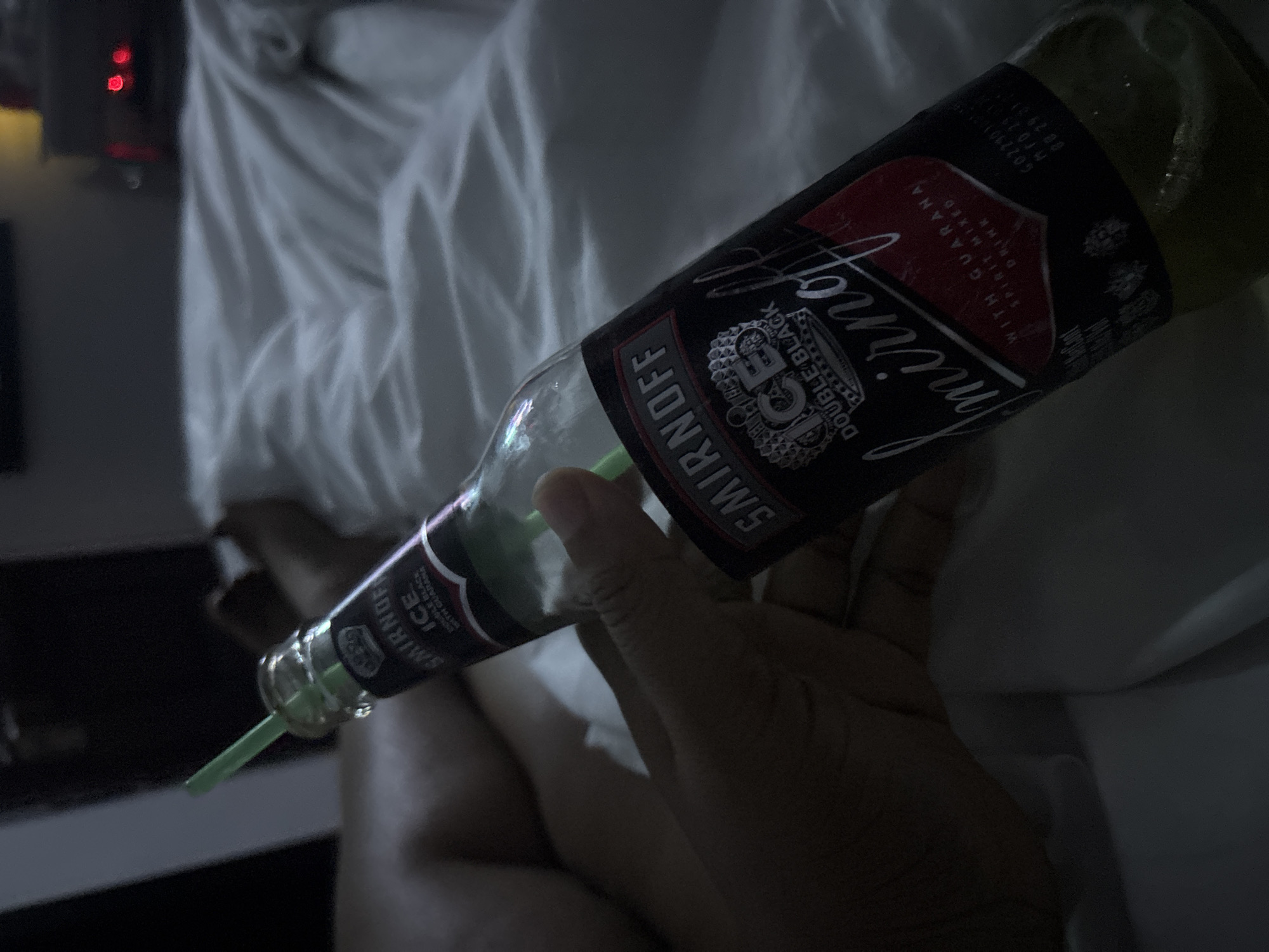 A hand holds a partially consumed bottle of Smirnoff Ice Double Black with Guarana, a pre-mixed alcoholic beverage. A green straw protrudes from the bottle. The background features a white, rumpled fabric and what appears to be a nightstand. The location is not discernible as Port Harcourt, Nigeria without additional information.