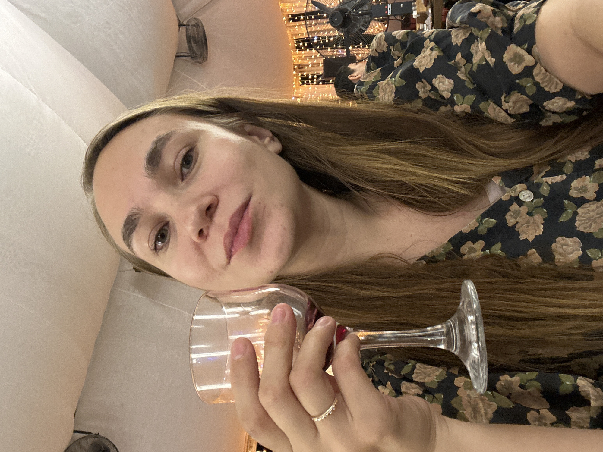 A woman is taking a selfie in a dimly lit indoor space, possibly a restaurant or event venue. She is holding a wine glass in her left hand, which has a delicate ring on her ring finger. She is wearing a dark dress with a floral pattern of light brown and green flowers. The background shows string lights and a large fan, suggesting a festive or possibly outdoor-covered setting. The woman's hair is long and brown, and she is looking directly at the camera with a slight smile. The overall lighting is warm and atmospheric. The location is not specified as Elista, Russia.