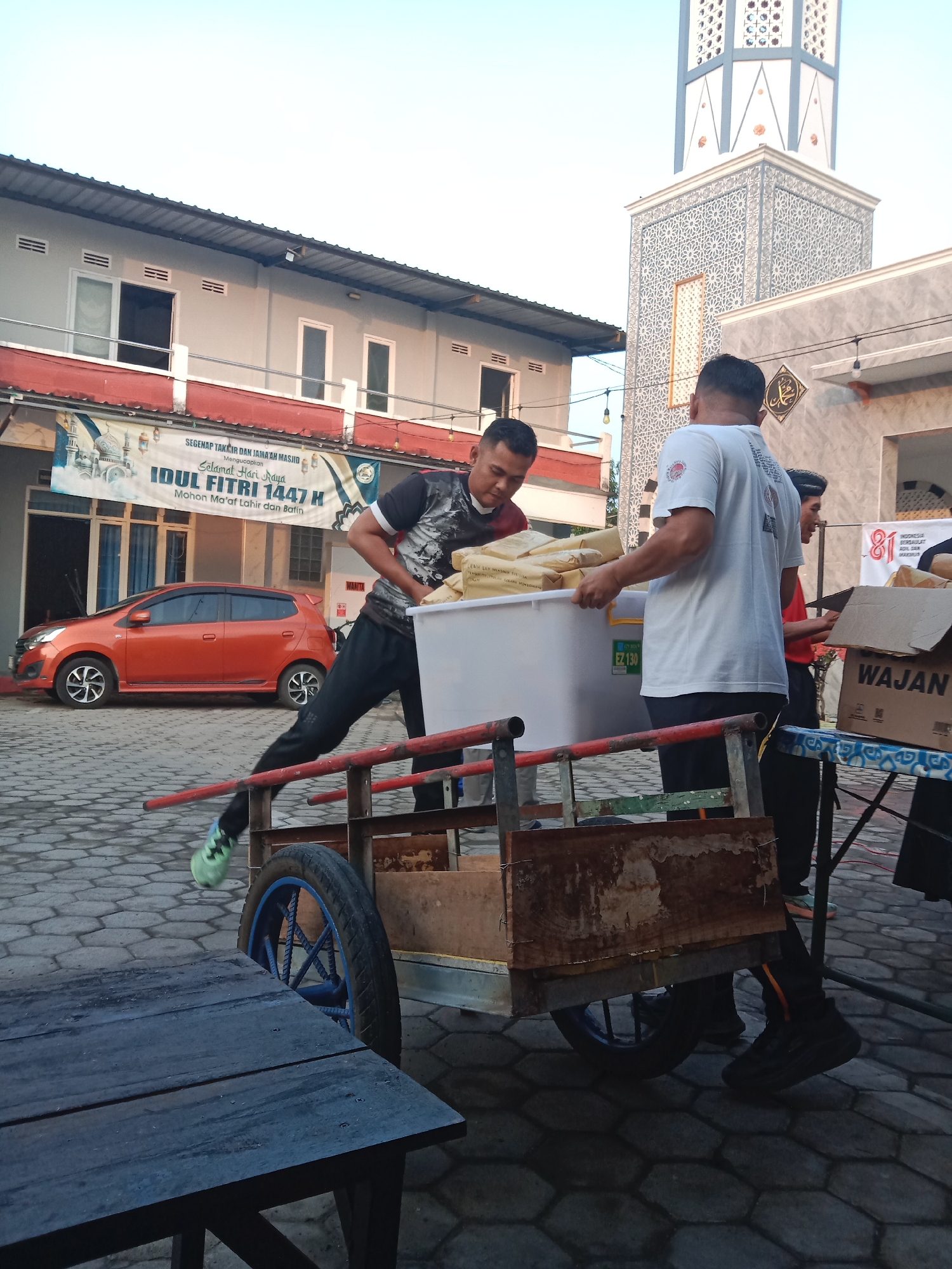 The image depicts an outdoor scene in Madiun, Indonesia, likely near a mosque, given the presence of a minaret and banner that reads "Segenap Takmir dan Jama'ah Masjid Mengucapkan Selamat Hari Raya Idul Fitri 1447 H". Two men are actively engaged in moving a large white plastic container filled with sacks, onto a rustic, wheeled cart. One man, wearing a black and grey patterned t-shirt and black pants with green athletic shoes, is pushing the cart forward. The other man, wearing a white t-shirt with graphic prints and black pants, is helping to maneuver the container. A third person, partially visible in a red shirt, stands behind them. In the background, an orange car is parked, and a building with multiple windows is visible. The ground is paved with hexagonal tiles. The lighting suggests it is either early morning or late afternoon.