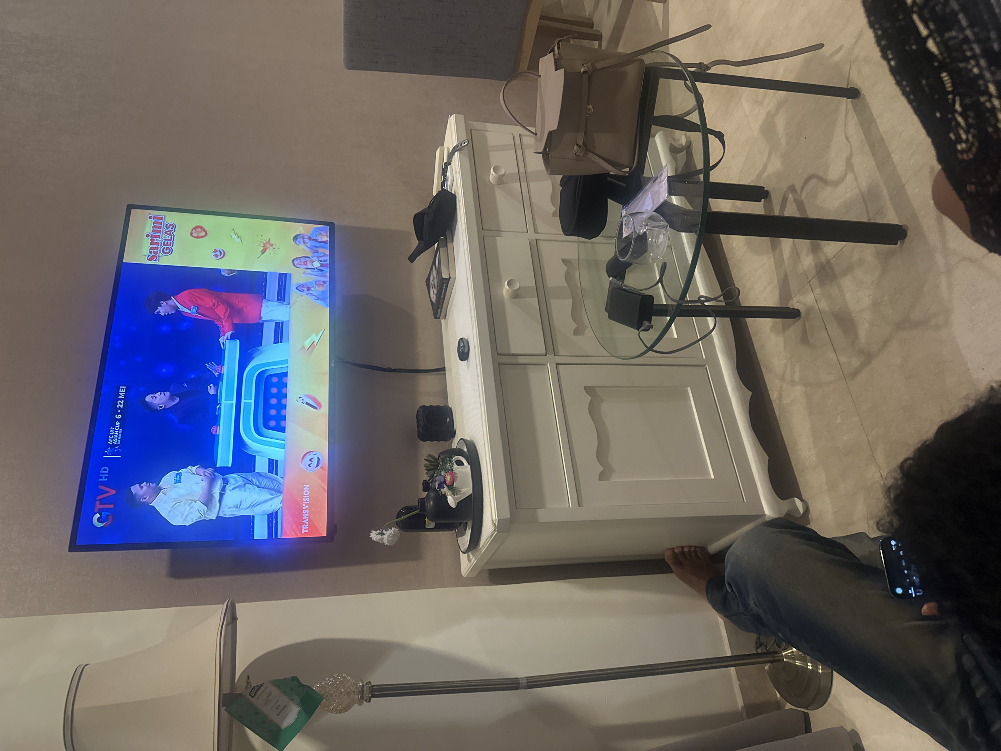 The image depicts an indoor scene in Central Jakarta, Indonesia. A television mounted on the wall displays the GTV HD channel, featuring the "AFC Cup Asian Cup 6-22 Mei" and the Transvision logo. Below the TV, a white chest of drawers is adorned with various objects, including a black hat, a framed picture, a Funko Pop figure, and a small vase with a flower. A round glass coffee table sits next to the chest of drawers, holding a tan handbag, a black beanie, and a pair of eyeglasses. To the left, there is a floor lamp with a green packet attached. A person wearing jeans is seated on the floor, holding a smartphone.