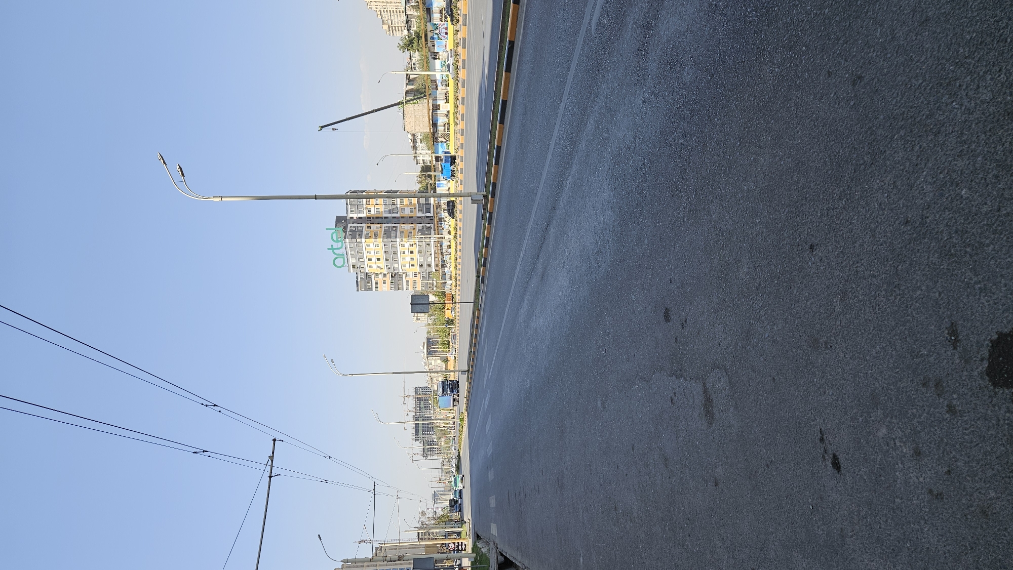 The image captures a view of a multi-lane road in Dushanbe, Tajikistan, likely taken during daytime given the clear sky and bright lighting. The road surface is asphalt, with distinct yellow and black markings delineating the lanes and sidewalks. In the background, a modern, tall building with the "artel" logo prominently displayed on its upper section stands out. Several other buildings, some appearing to be under construction with cranes visible, line the street. Vehicles, including cars and a truck, are present on the road, suggesting ongoing traffic. Streetlights are positioned along the roadside, and overhead electrical wires, possibly for public transportation, crisscross the sky. The scene depicts an urban environment with a mix of existing infrastructure and ongoing development.