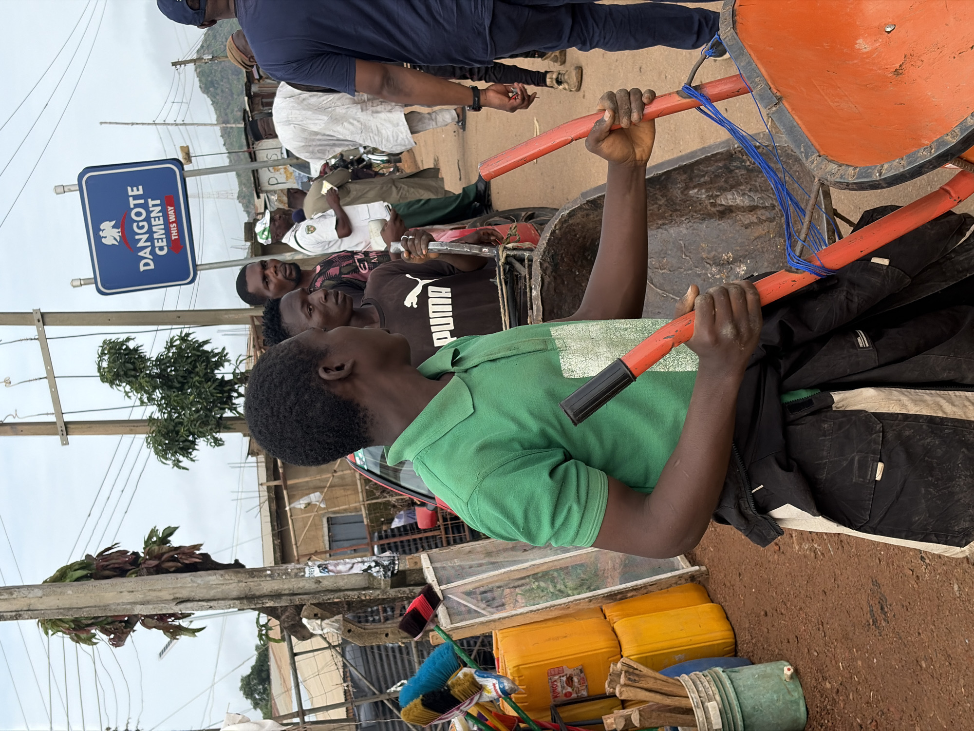 A group of people are gathered outdoors in a dusty urban environment, possibly in Gura Top, Nigeria, near a Dangote Cement sign. In the foreground, a young Black male wearing a green polo shirt and black pants is holding the handles of a bright orange wheelbarrow. He is looking to his left, away from the camera. Behind him, other individuals are visible. One man in a black t-shirt with a white Puma logo is looking at the camera. Another man in a white shirt and cap is standing with his back to the camera, and others are further in the background. The setting appears to be a street or market, with various objects including yellow plastic containers, brushes, and wooden sticks scattered around. Electrical wires are strung across poles overhead, and some buildings are visible in the background. The overall impression is one of a busy, outdoor commercial area.