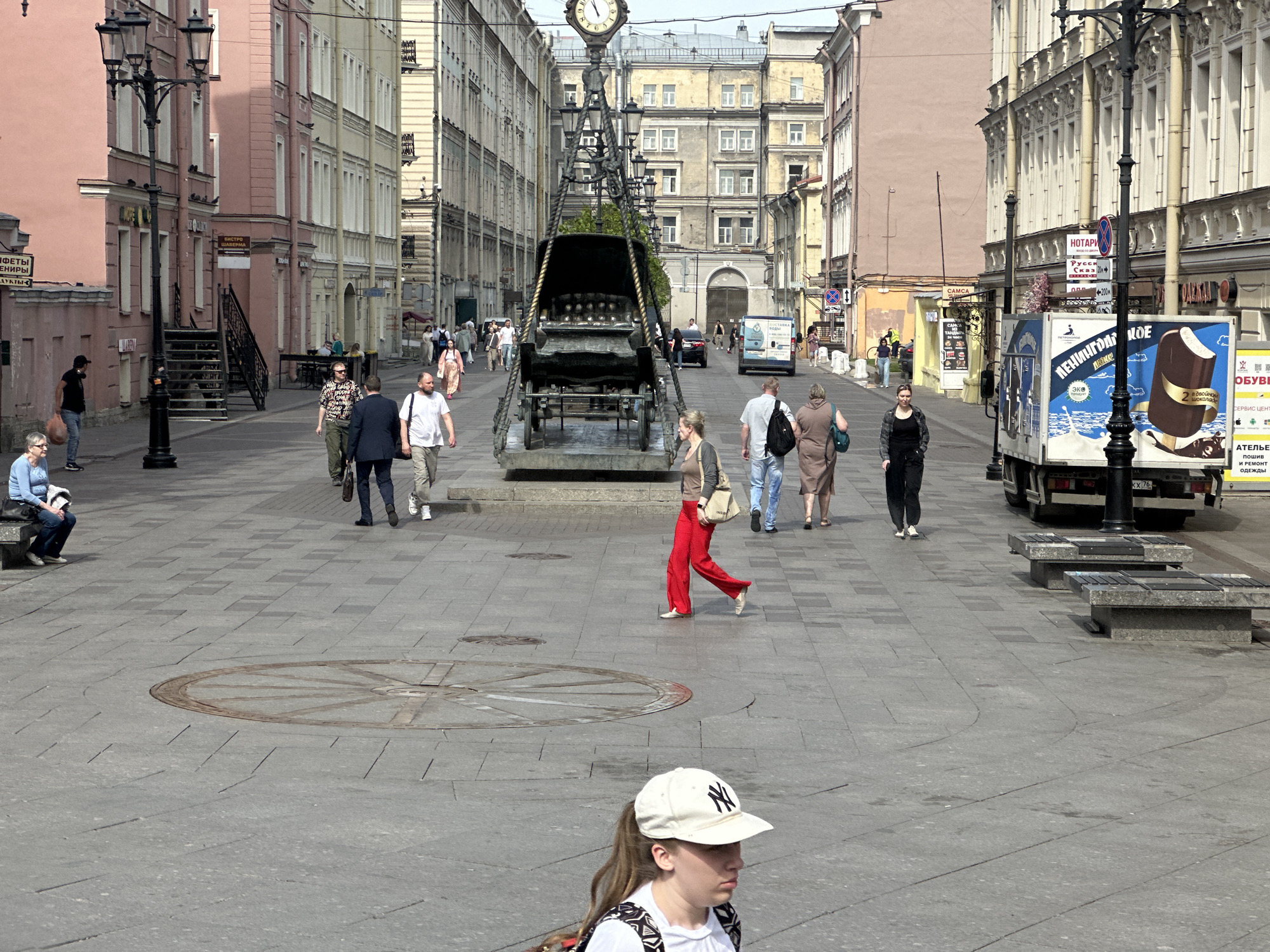 The image captures a street scene in Saint Petersburg, Russia, on a sunny day. The scene features a wide pedestrian zone paved with gray tiles, with several people walking around. There is a unique sculpture of a horse-drawn carriage raised up by ropes, hanging from a clock tower in the middle of the street. In the foreground, a young girl wearing a white New York Yankees baseball cap walks towards the camera. A woman in red pants is walking near the sculpture. Several other people are dispersed in the background, walking in various directions. A truck with ice cream is parked on the right side of the street. Buildings line both sides of the street, varying in color and architectural style. Street lamps are visible along the sides of the pedestrian zone.