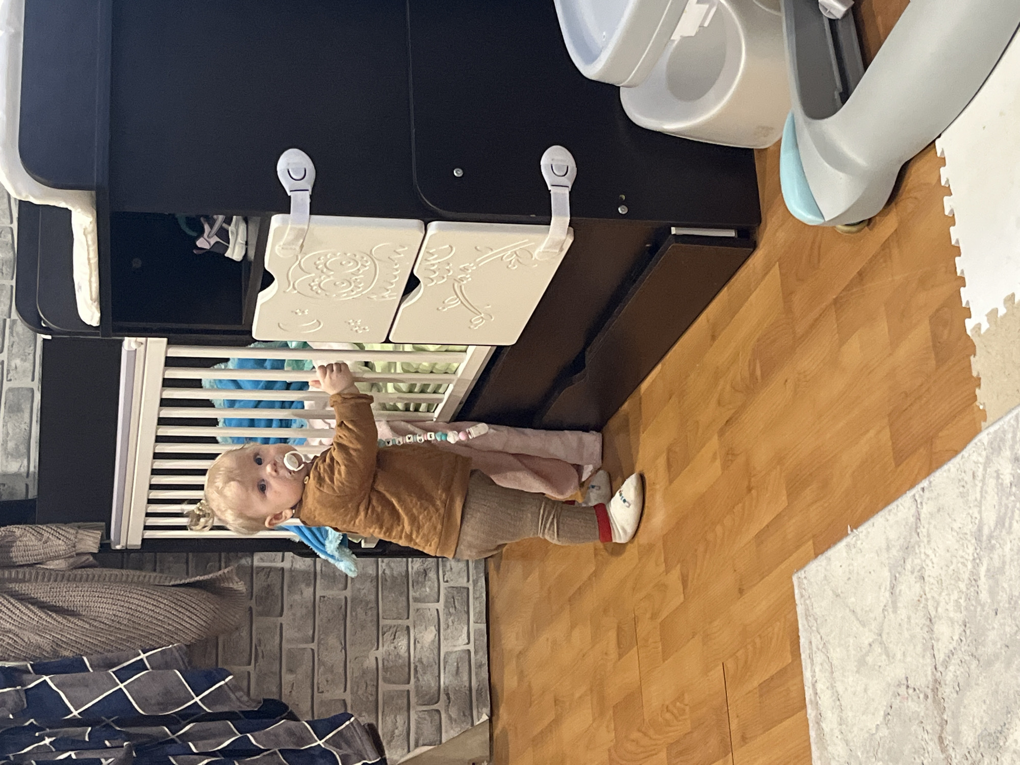 This image, captured in Vesyoloye, Russia, depicts an indoor domestic scene, likely a child's room or play area. The primary subject is a young child, appearing to be a toddler with light blonde hair and blue eyes, wearing a brown long-sleeved top, light brown pants, and white socks with a red band. A white pacifier is in the child's mouth, attached by a beaded clip.
The child is standing upright, holding onto the white slatted rail of a baby crib or cot with their left hand, and looking directly at the camera with a neutral, curious expression. Inside the crib, a blue plush toy and other soft items are visible. Behind the crib is a dark brown cabinet or storage unit, to which two white drawer fronts with embossed decorative patterns are attached. These drawers are secured with white child safety latches, indicating a child-proofed environment.
The flooring consists of light brown wooden laminate or parquet, partially covered by a white, interlocking soft play mat in the upper right of the frame and a shaggy white rug in the bottom right. A portion of the wall to the left features a gray brick-patterned wallpaper or paneling. In the upper right corner, part of a light gray and white high chair or similar baby seat with a light blue accent is visible. Other textiles, including a blue, gray, and white patterned blanket and a textured brown/beige blanket, are seen in the lower left. The image is oriented sideways (rotated 90 degrees clockwise). No visible text can be clearly discerned.