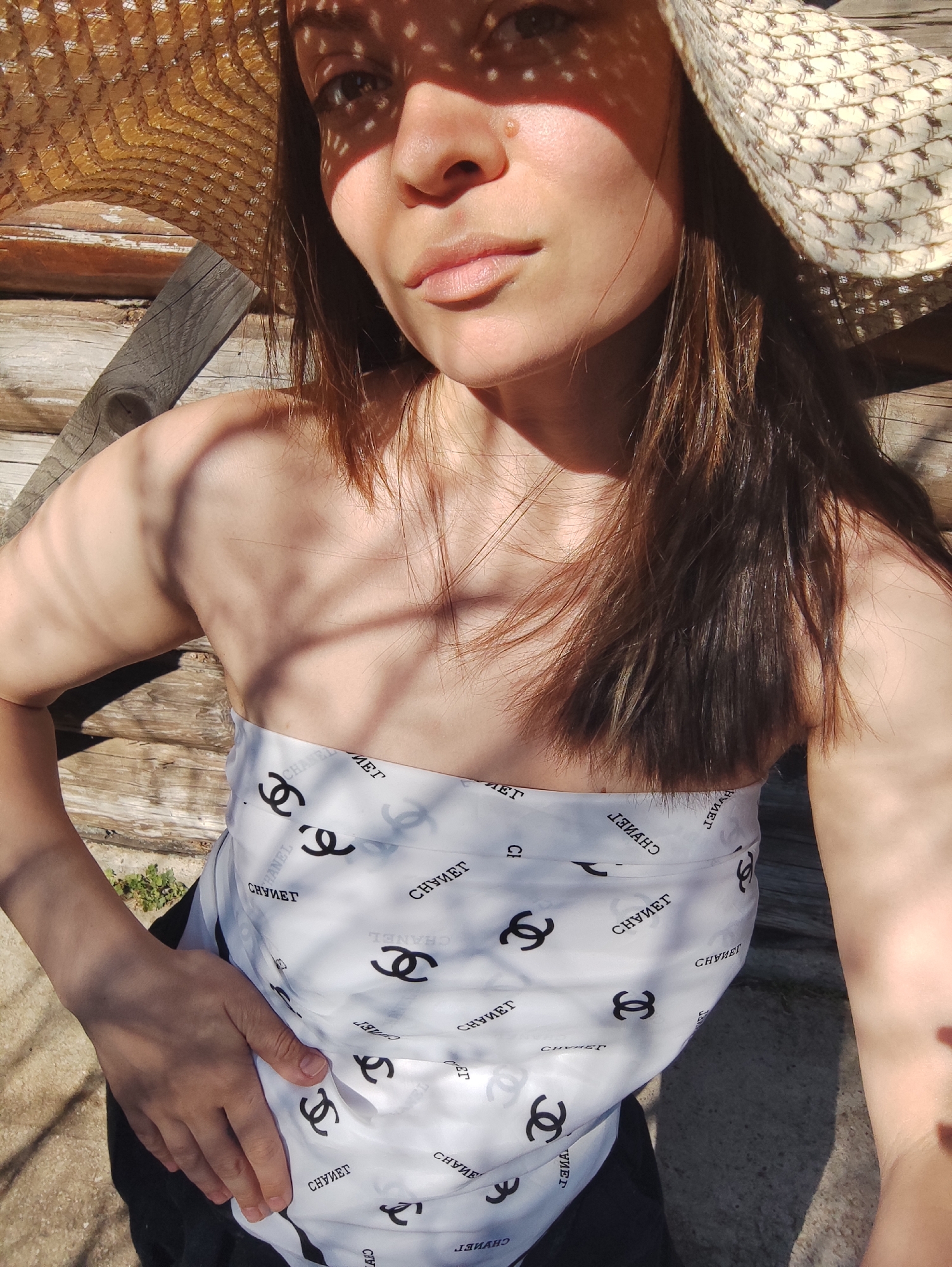 A young woman is depicted in a sunlit outdoor selfie, positioned in front of a rustic wooden wall made of logs. She wears a wide-brimmed straw hat that casts intricate, dappled shadows across her face and shoulders, indicating strong, direct sunlight. Her long, dark hair frames her face and shoulders. She is wearing a white strapless top or bandana, patterned with black repeating motifs. These motifs include the interlocking 'C' logo, strongly associated with Chanel, and various textual elements. The OCR identifies these as "CHANE", "HANEL", "СНУИЕГ", "МЕГ", "ДЕГ", "CHANEL", "CH", "CHE", "8", and "УИЕГ", with some text appearing stylized or inverted. The woman's expression is neutral, with slightly parted lips. Her right hand is visible, resting on her waist or hip. The scene conveys a bright, clear day in what appears to be a rural or natural setting, aligning with the location context of Фоки, Russia.