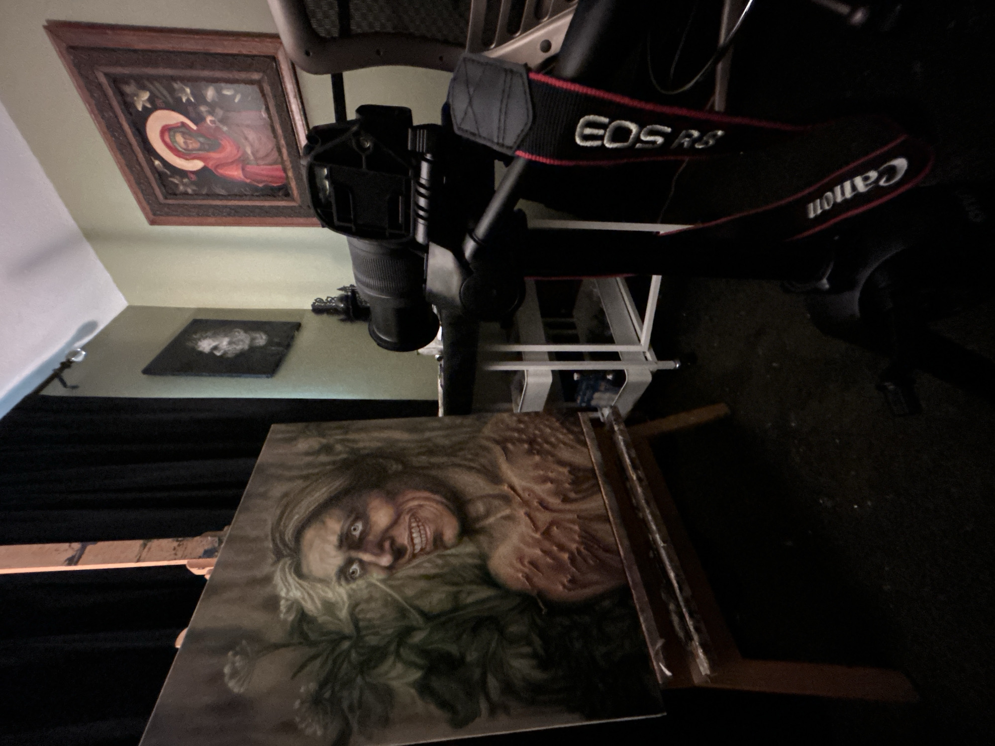 This media file captures an indoor scene, likely an art studio or a dedicated space for art documentation, located in Cape Coral, United States of America.
In the foreground, a striking and unsettling painting is prominently displayed on a wooden easel, positioned upright. This artwork depicts a grotesque figure with wide, staring eyes, a contorted face, and a baring, toothy grin, evoking a sense of unease or horror. The figure has long, light-colored hair and visible textured details on the body, set against a dark, possibly organic or floral background.
Dominating the mid-ground, a professional camera setup is visible, angled across the frame. A black Canon mirrorless camera, identified by "EOS R5" and "Canon" printed on its red and black strap, is mounted on what appears to be a tripod or overhead arm, aimed downwards towards the painting on the easel. The camera's lens is clearly visible.
In the upper left portion of the image, hanging on a light green wall, is another framed artwork. This piece appears to be a religious icon, possibly depicting the Virgin Mary or a saint, characterized by a halo, traditional drapery, and a serene expression. Below this, on a darker surface, a smaller, dark-framed artwork features a lighter, abstract or cloud-like shape.
The overall lighting in the scene is dim, suggesting either a controlled lighting environment for photography or simply a low-light interior. The background behind the camera setup is mostly dark and indistinct, possibly consisting of dark fabrics or wall coverings. The scene indicates the activity of photographing artwork, likely for documentation or promotional purposes.