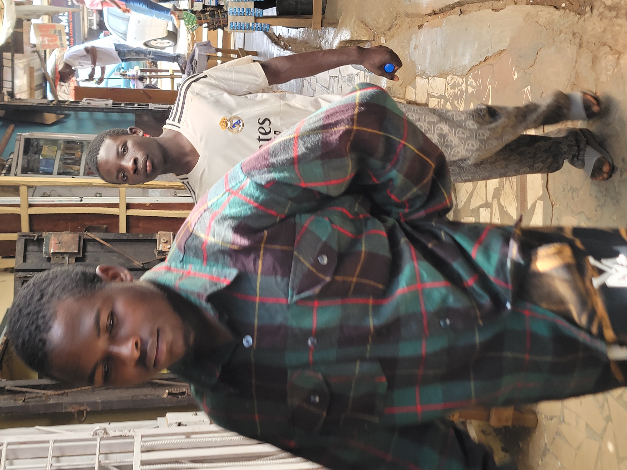 Two young men are in an outdoor setting that appears to be in Jos, Nigeria. One man is closer to the camera, wearing a green plaid shirt. The other man is further away, wearing a white Real Madrid soccer jersey and grey patterned pants. He holds a small blue object. The location includes a building with a window display behind the man in the jersey and a white painted wall in the background. Pedestrians are visible further back in the scene.