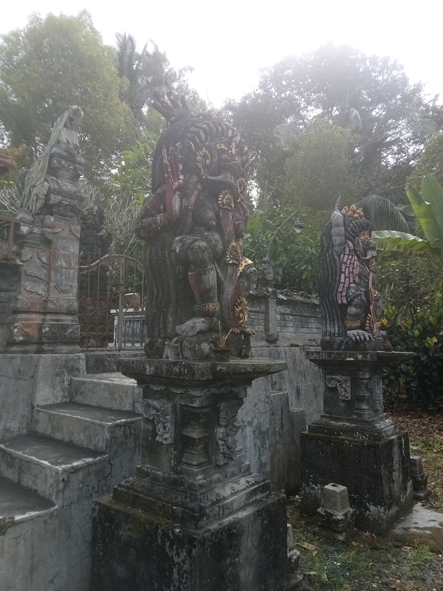 Two stone statues, likely of mythological figures, stand on pedestals in an outdoor setting with lush green foliage and trees in the background. The statue on the left is partially obscured by foliage and features a veiled head. The statue in the center is dark and imposing, with intricate carvings and what appear to be multiple arms. The statue on the right is also dark, with pink accents and a detailed headdress. Concrete steps lead up to the left of the statues, and a rusted metal gate is visible behind the central statue. The ground around the statues is covered in dry leaves and grass. The overall scene suggests a temple or an ancient site, possibly in a tropical climate.