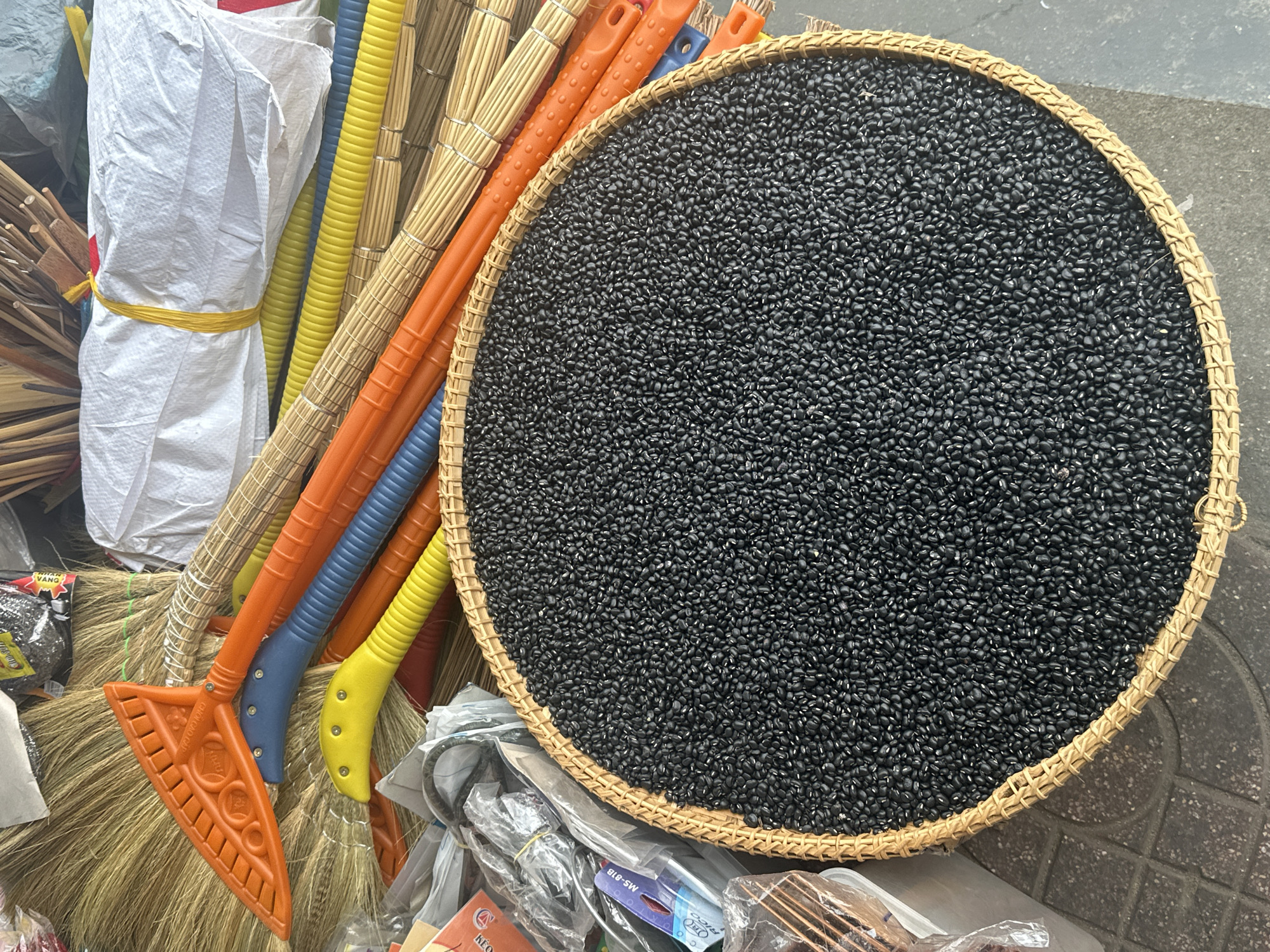 The image shows an outdoor scene in Bac Nha Trang Ward, Vietnam. A round, woven basket is filled with a layer of small, black seeds. To the left of the basket are several brooms with multi-colored plastic handles (orange, blue, yellow) and straw bristles. There is also a white, tied-up bag and other miscellaneous items in plastic packaging in the foreground. The surface the items are resting on appears to be concrete with a tile pattern visible in the lower right corner.