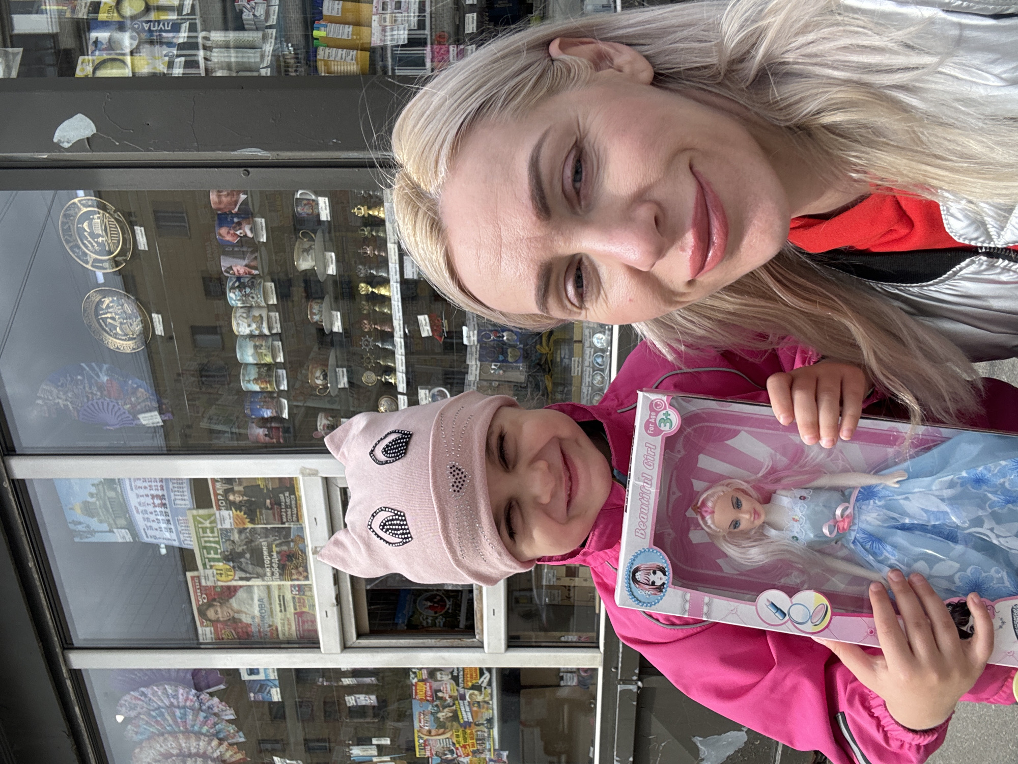 A fair-skinned woman with blonde hair and a younger child are pictured outside of a storefront in Saint Petersburg, Russia. The woman is wearing a silver jacket over a red shirt. The child is wearing a pink jacket and a pink hat with decorative eye-like embellishments. The child is holding a boxed doll. The storefront has various items displayed in the windows, including decorative plates, mugs, and periodicals. The items on display and in view in the window indicate a location in Saint Petersburg, Russia.