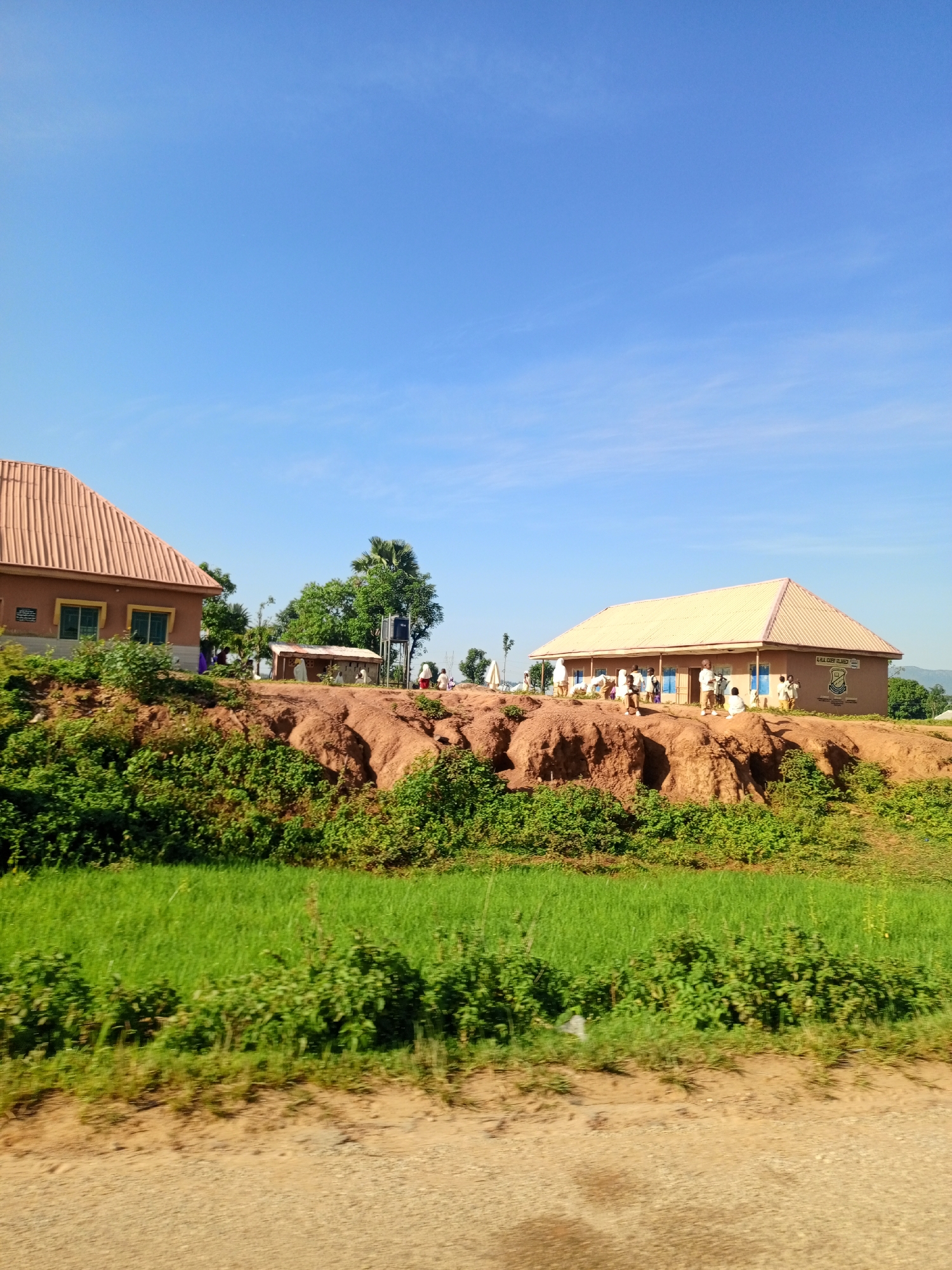 The image depicts a scene in the city of Rinjim Gani, Nigeria. In the foreground, a dirt road runs horizontally. Behind the road, lush green grass is visible. The middle ground is dominated by a reddish-brown, eroded hillside. On top of this hill, two school buildings are situated. The building on the left has an orange, tiled roof and light brown walls with green-framed windows. The building on the right, larger and longer, also has an orange, tiled roof and light brown walls. This building has blue-framed doors and windows, and a crest is visible on its side with text that reads "AL-HAFIDH ADENIYI GBADEGESIN". Several people, appearing to be students in uniforms, are gathered outside and around the buildings. Some are standing in small groups, while others are seated. Behind the school buildings, trees and vegetation can be seen. The sky is a clear, bright blue with a few wispy clouds. The overall impression is of an educational institution in a rural or semi-rural setting.