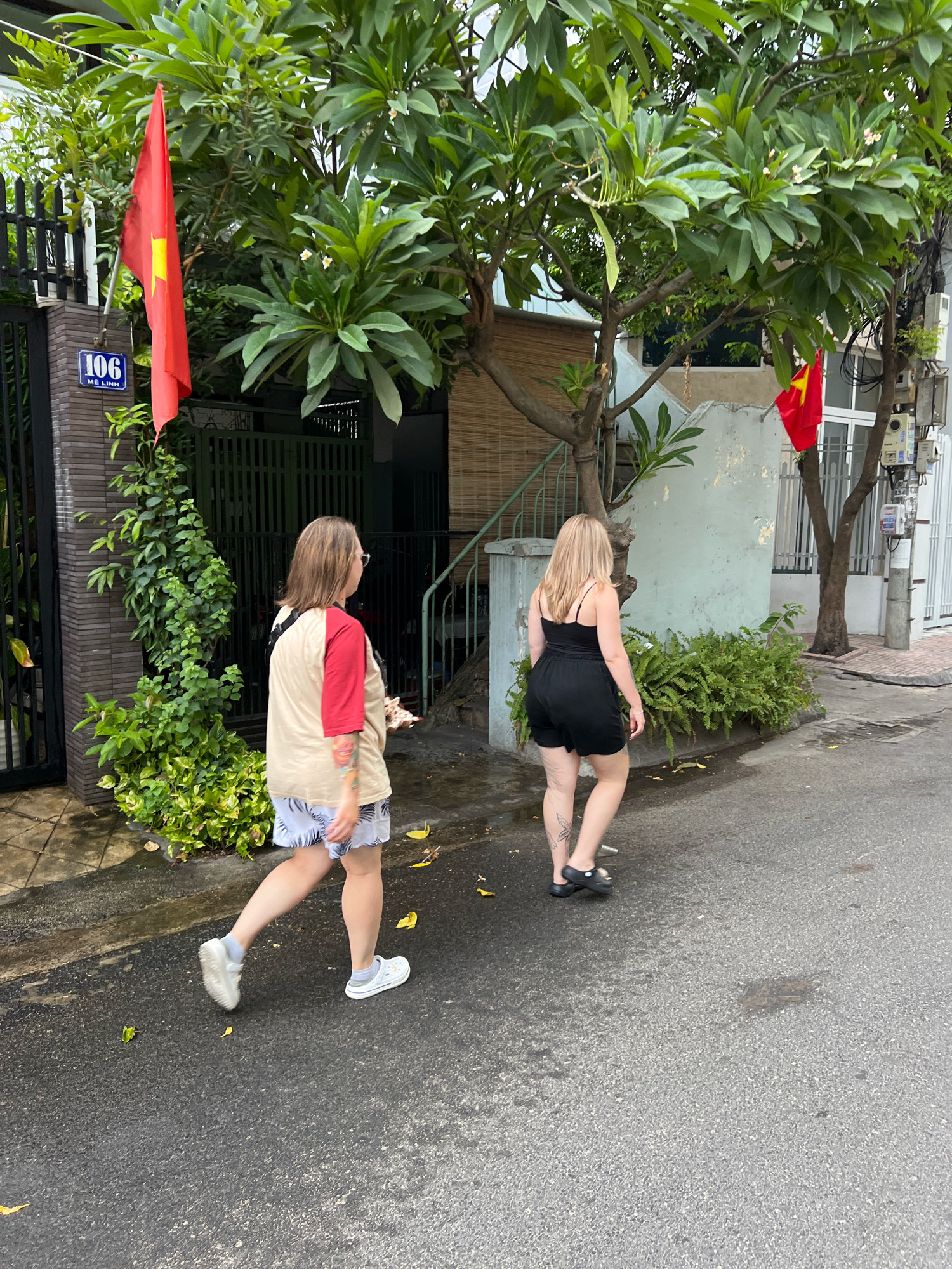 The image depicts a street scene in Nha Trang, Vietnam, characterized by lush greenery and urban infrastructure. Two individuals, both women, are walking away from the camera on a wet asphalt road.
The woman on the left has shoulder-length brown hair and wears glasses. Her attire consists of a cream-colored t-shirt with red sleeves and patterned shorts. She is carrying a bag over her shoulder, and her left arm is adorned with colorful tattoos. She is wearing white sneakers.
The woman on the right has long blonde hair and is dressed in a black tank top and black shorts. She is wearing black slip-on shoes. A butterfly tattoo is visible on her left ankle.
The environment includes a building with a textured wall, a gate, and a fence. A red flag with a yellow star, likely the Vietnamese flag, is prominently displayed on a flagpole. Another similar flag is visible in the background. Trees with dense green foliage are scattered throughout the scene, and several plants, including ferns, grow along the side of the building. The ground is damp, suggesting recent rain. Electrical conduits and meters are attached to a pole in the background, indicating the presence of urban utilities. A house number "106" with Vietnamese text below it is affixed to the building on the left.