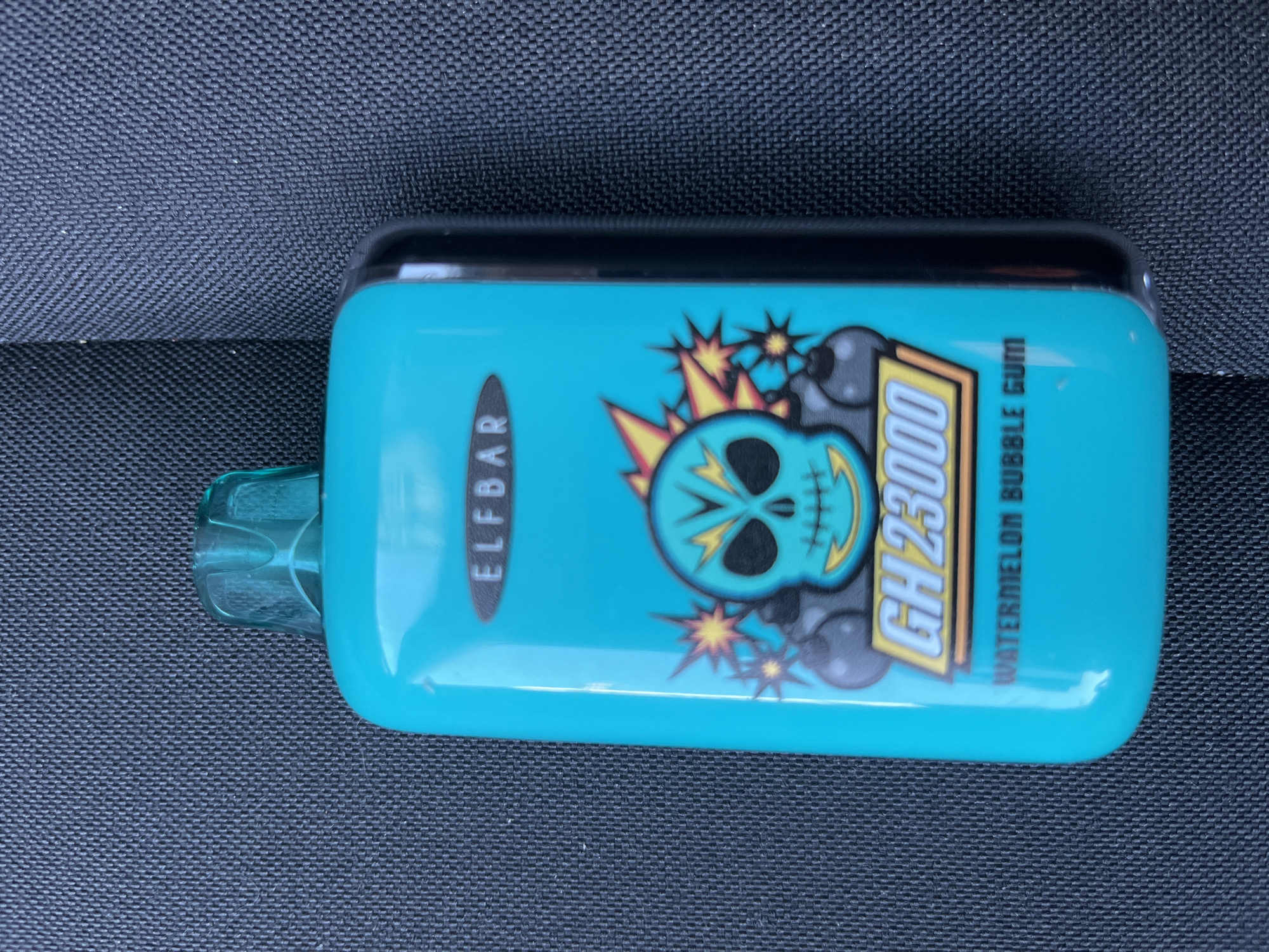 The image shows a bright teal disposable electronic cigarette device resting on a dark, textured surface, likely fabric. The device is rectangular with rounded edges and features a black top and a translucent teal mouthpiece extending from the left side.
Prominently displayed on the device is a graphic of a stylized skull with a turquoise face, black eyes, and teeth. Explosions with yellow and orange starbursts emanate from behind the skull. To the right of the skull, in a yellow rectangular box with a black outline, is the text "GH 23000". To the left of the skull, vertically oriented within a black oval shape, is the text "ELFBAR". Further to the right, also vertically oriented, is the text "WATERMELON BUBBLE GUM".
The background is a uniform dark color with a distinct woven texture, suggesting it is a piece of furniture or upholstery. The lighting appears to be natural daylight, casting a soft, even illumination on the object. No people are visible in the image. The scene is static, with the electronic cigarette as the sole subject. The location is identified as Kharkiv, Ukraine, though no specific visual cues within the image confirm this. The overall impression is a close-up product shot.