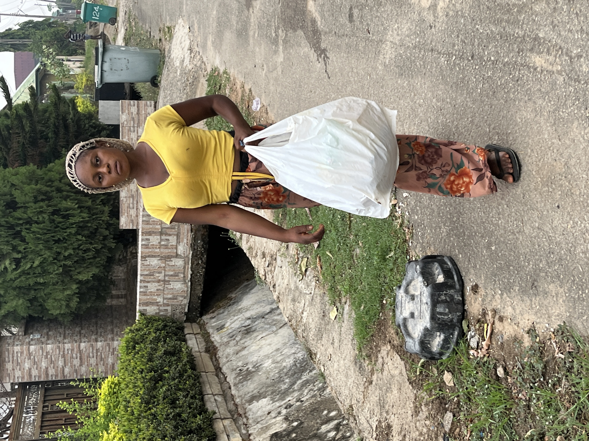 A woman wearing a yellow shirt, floral pants, and sandals stands on a paved surface next to a grassy curb. She is holding a white plastic bag. Next to the curb is a black plastic object. A drain runs along the edge of the pavement. Behind the woman are a building, bushes, and a trash can. The scene is located in Abuja, Nigeria.