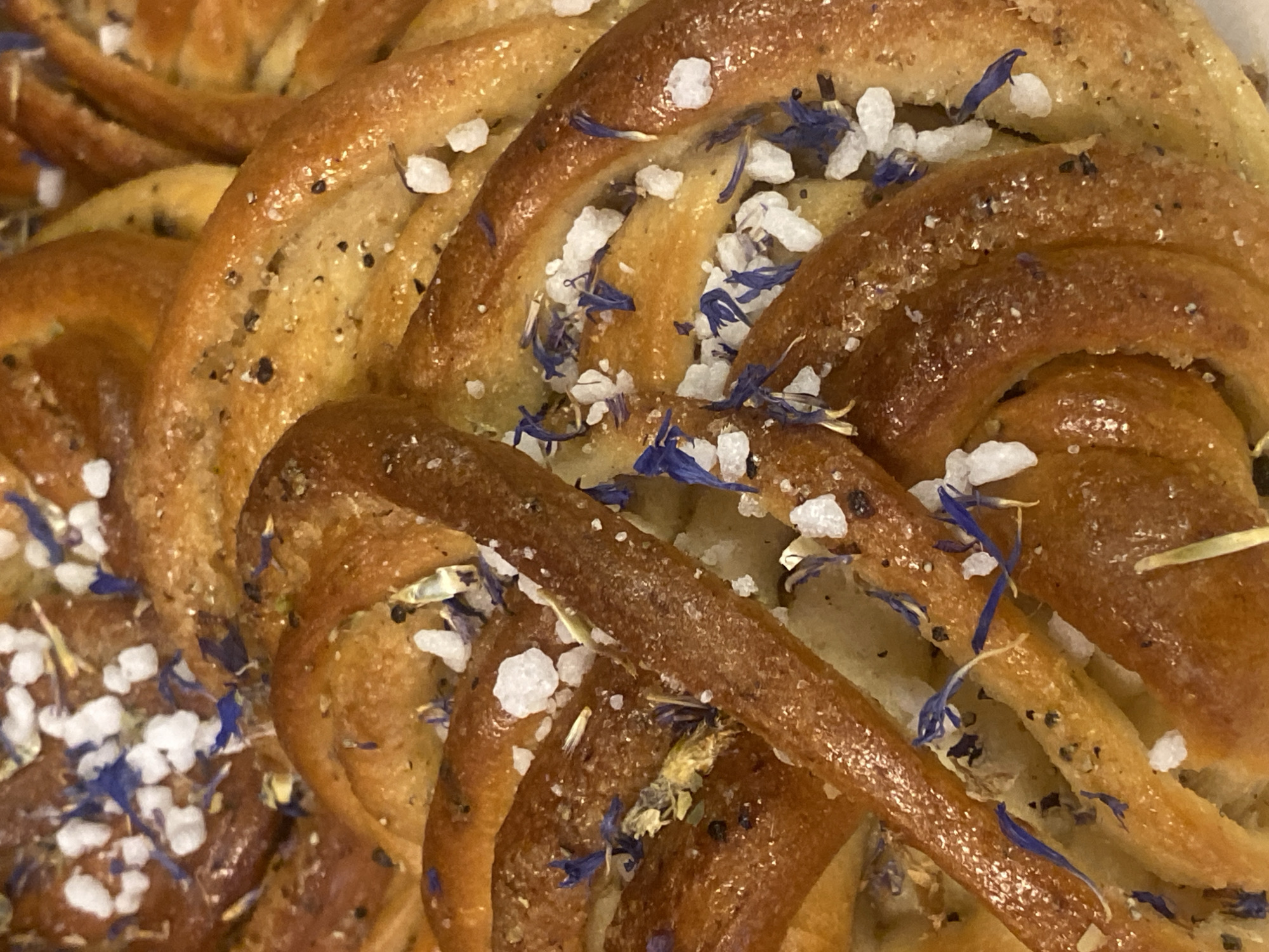 This is a close-up, overhead shot of a braided pastry, likely a type of sweet bread. The pastry has a rich, golden-brown crust with glistening highlights, indicating it may have been brushed with an egg wash or syrup. It is generously sprinkled with coarse white sugar crystals and small, dark specks that could be poppy seeds or other spices. Scattered amongst the sugar are delicate, deep blue petals, likely from cornflowers, adding a decorative touch and a hint of floral aroma. The pastry is intricately twisted, revealing layers and a soft interior. The lighting is warm and even, suggesting an indoor setting, possibly a bakery or kitchen. There are no people visible in the frame. The scene is focused solely on the textures and details of the baked good, emphasizing its appetizing appearance. The context of Warsaw, Poland, suggests this could be a traditional Polish pastry. There is no visible text in the image.