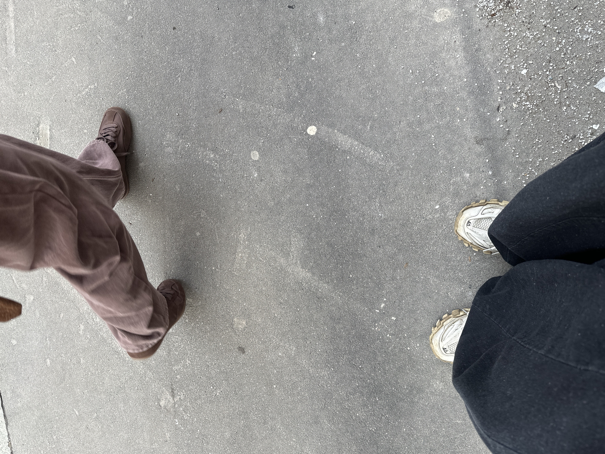A photograph from an elevated, first-person perspective captures two individuals standing on a paved surface, likely a sidewalk.
The individual on the left wears light brown corduroy pants and brown shoes. The individual on the right is clad in black pants and white, dirty sneakers.
The background is the gray paved surface. Some small, light colored rocks can be seen on the right side of the surface.
The location is Moscow, Russia.