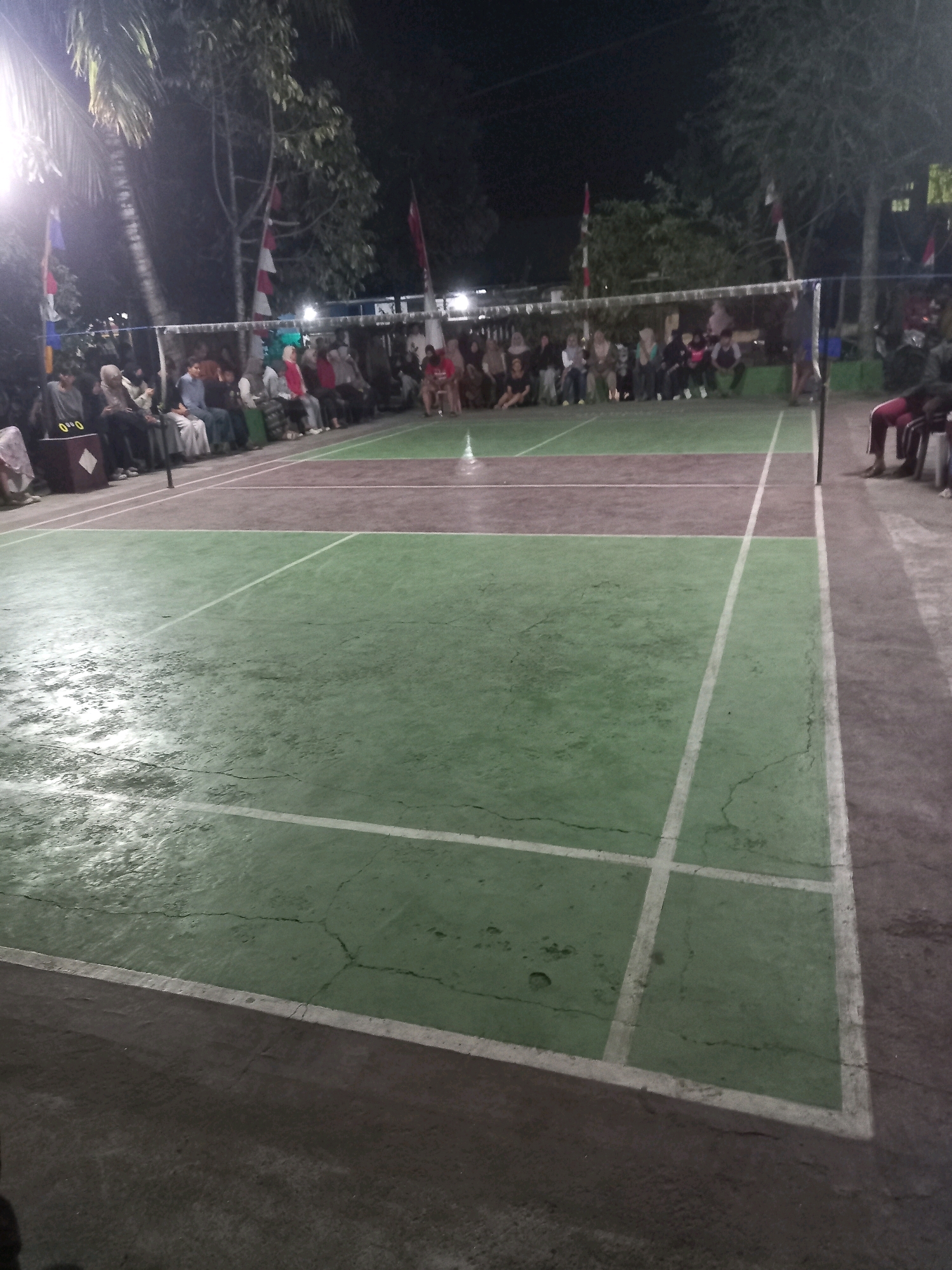 This is a nighttime photograph of an outdoor badminton court in Banyumulek, Indonesia. The court has a green surface with white lines marking the boundaries. A net divides the court in half. Spectators are seated on benches to the left and right of the court. They appear to be watching a game or event. The lighting is provided by artificial lights, which cast a bright glow on the court. The overall atmosphere is that of a community gathering or sporting event.