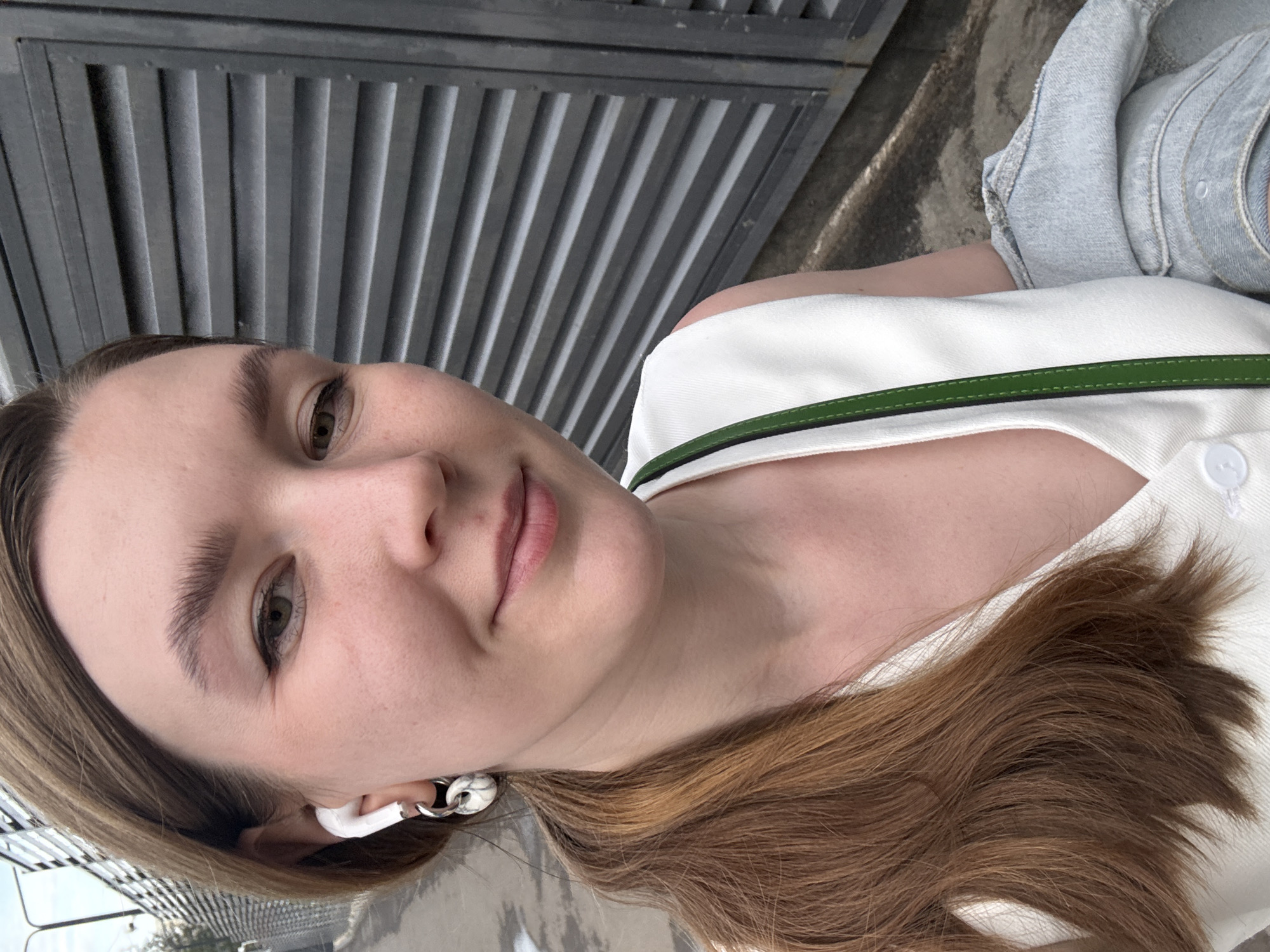 A young woman is shown in a close-up, angled shot. She has fair skin, light brown hair, and is wearing a white top with a green strap. She is wearing what appear to be white wireless earbuds. The background consists of a metal fence and a glimpse of a city scene, consistent with being in the city of Izhevsk, Russia.
