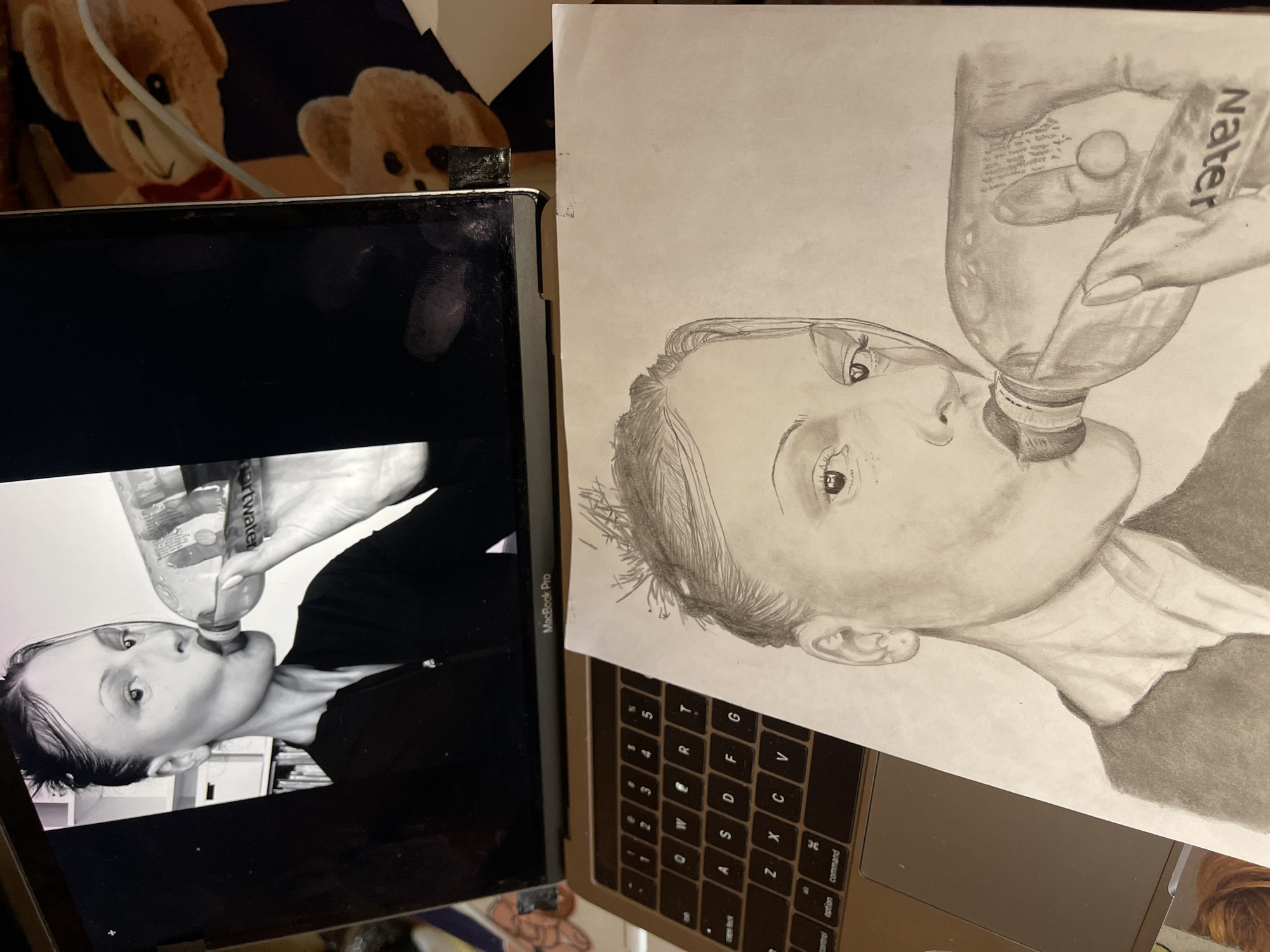 An elevated view captures an indoor scene in Jos, Nigeria, featuring a silver Apple MacBook Pro laptop and a pencil drawing. The laptop's screen displays a monochrome photograph of a person, likely female, with slicked-back hair, looking rightward while drinking from a plastic bottle labeled "SmartWater." The person wears a dark, collared garment with a partial zipper visible. The background of the photograph is blurred, showing light-colored rectangular forms on the left. Positioned on the laptop's keyboard and extending onto the trackpad, a monochrome pencil drawing replicates the subject from the screen, rendered in graphite shades on white paper. The drawing details the individual's face, neck, shoulder, and hand gripping the bottle, with the inverted "Water" label clearly visible. To the right of the laptop, a blurred fabric surface patterned with repeated light brown teddy bears on a dark background is visible. A thin white cable crosses this background from the upper right.
