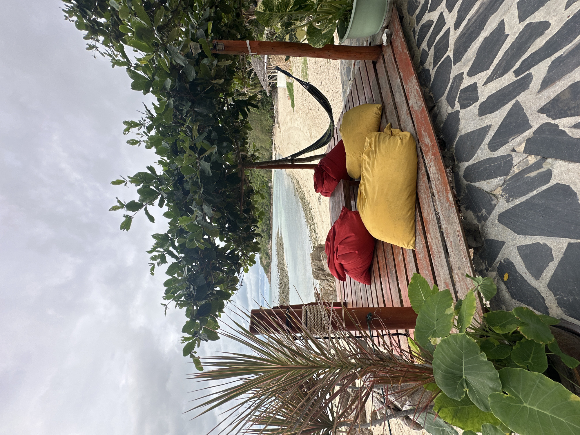 This is a photograph taken outdoors in the Ko Pha-ngan District, Thailand. The scene is a wooden deck situated near a beach. On the deck are several large, colorful floor cushions, two red and two yellow. A hammock is strung up between two wooden posts, partially shaded by the leaves of a large tree. Beyond the deck, a sandy beach meets a body of calm, light-colored water. The sky is overcast with gray clouds. In the foreground, a potted plant with large green leaves and a spiky plant with reddish-green leaves are visible. The ground in the foreground is paved with irregular, dark gray and light gray stones.