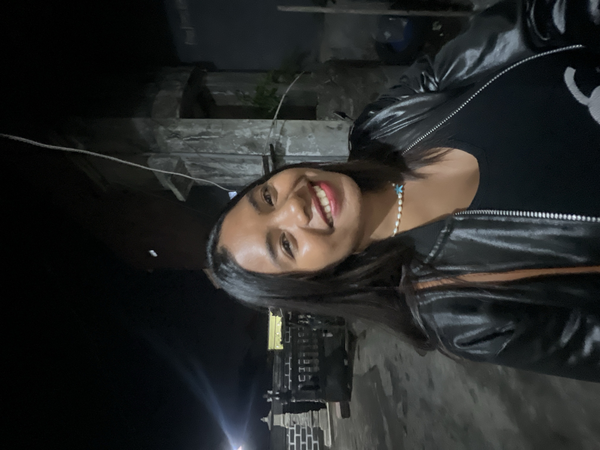 A young woman with dark hair and fair skin smiles at the camera in this medium shot. She is wearing a black leather jacket over a black tank top, and a pearl necklace with a blue starfish pendant. Her makeup includes red lipstick and eyeliner. The background is dark, suggesting it is nighttime. The location is a street in Denpasar, Indonesia, with a glimpse of traditional Balinese architecture in the background, specifically a stone structure with carvings and a patterned wall. There is also a utility pole with wires and some foliage visible. The ground appears to be paved and wet, reflecting the available light.