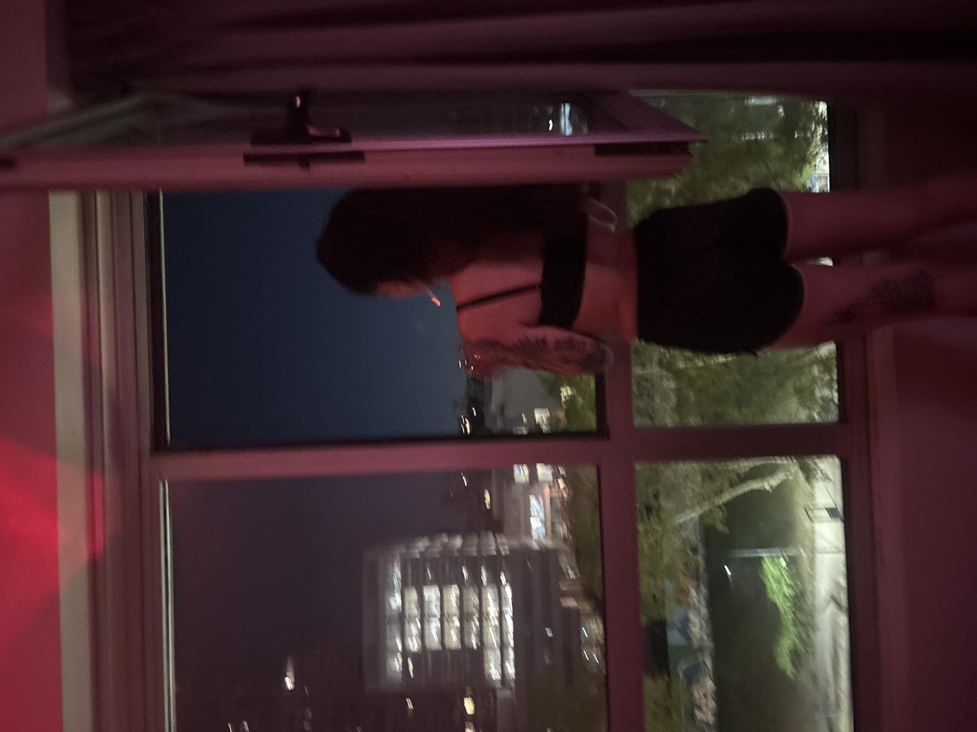 A woman in a black top and dark shorts stands in an open window, smoking. Her left leg is visible, with tattoos on her thigh. Outside the window, a cityscape is visible at night. The buildings appear illuminated, suggesting an urban environment. The image is taken from inside a room with red walls and a partial view of curtains. The location is likely Buenos Aires, Argentina, based on the architectural style and urban density visible through the window.
