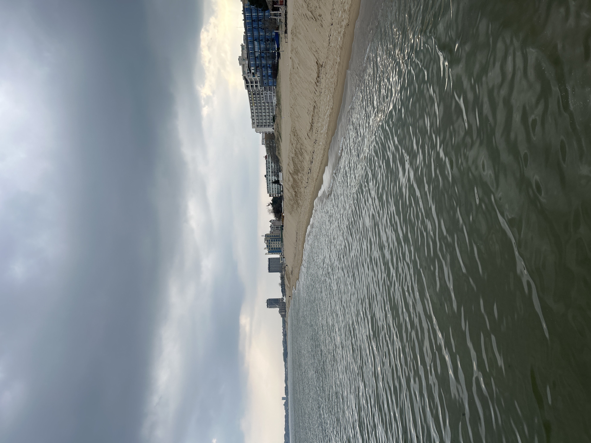 The image presents a coastal scene on an overcast day. The right side of the frame is dominated by rippling water, suggesting a sea or ocean. A narrow strip of sandy beach borders the water. Along the beach are several buildings, including what appears to be hotels or apartments, stacked vertically from top to bottom along the left side of the coastline. The sky is heavily clouded, indicating potentially stormy weather. The image is oriented with the sky and clouds along the left side, the water on the right, and the buildings stacked on the upper-left to lower-center of the frame.