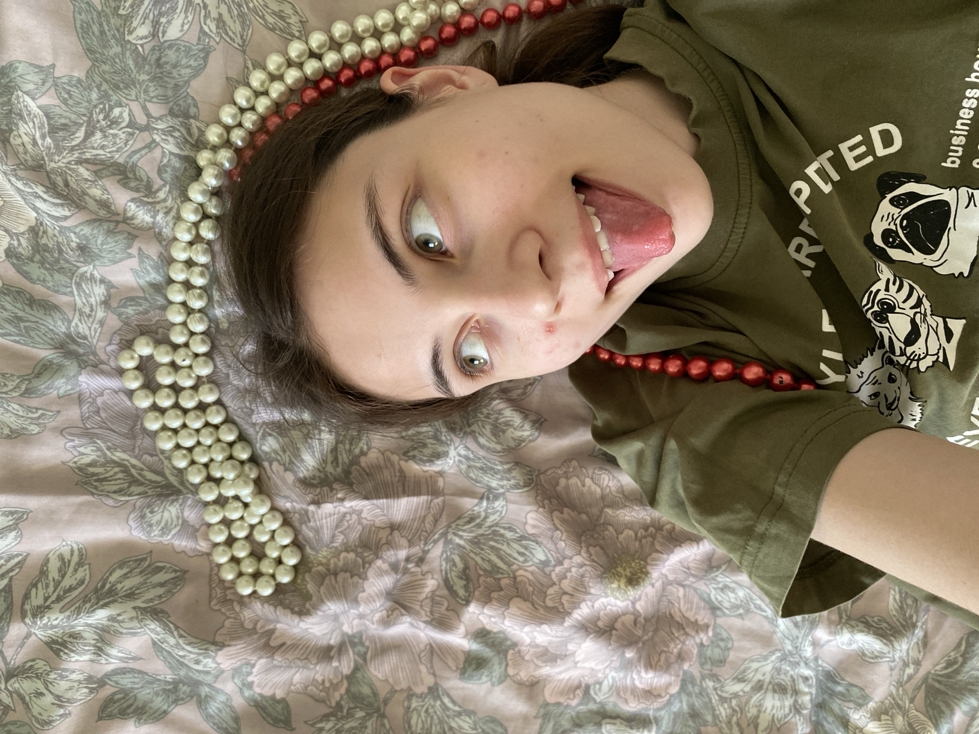A young woman is lying on a floral patterned surface. She is wearing an olive green T-shirt with graphic prints and text. Strands of white and red beads are draped around her head. Her mouth is open, sticking her tongue out and her eyes are wide. There is no information to identify the location in Votkinsk, Russia.