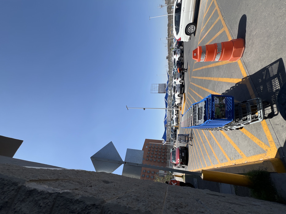This image depicts an outdoor scene at a commercial parking lot in León, Mexico, under a clear blue sky. In the immediate foreground, a stack of approximately 10-12 blue shopping carts is visible on a cart return bay, positioned on concrete paving marked with yellow diagonal lines. To the right of the carts is a large orange traffic cone with two white reflective bands. On the far left, a textured grey concrete wall or pillar partially frames the shot, with a yellow bollard and a portion of a person wearing an orange vest visible near its base.
In the mid-ground, a red SUV is parked near another cart return bay. Further into the parking lot, numerous vehicles, predominantly white and light-colored SUVs and sedans, are parked beneath blue fabric shade structures. One individual wearing a dark uniform is visible near these parked vehicles, appearing to bend over or interact with the ground or a vehicle. In the far distance, a tall, multi-story building with reddish-brown facade and numerous windows is visible on the left. Several streetlights punctuate the scene, and a large metal billboard structure is discernible in the background on the right, beyond the shaded parking area. The ground consists of asphalt with yellow painted lines for parking demarcation.