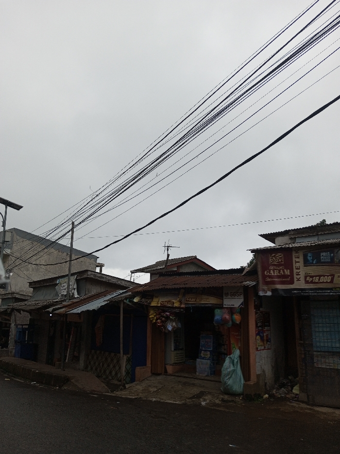 The image presents a street scene in Pacet, Indonesia, captured under a uniformly overcast sky. The foreground features a dark asphalt road, bordered on the left by a low, weathered concrete curb. Extending along the right side of the road is a row of contiguous, one-story commercial structures or residences, characterized by corrugated metal roofs, some exhibiting rust. Multiple black electrical and telecommunication wires are densely strung across the upper portion of the frame, originating from out-of-frame poles and a visible utility pole on the far left, which is equipped with a solar panel array.
No individuals are visible within the frame. The leftmost structure appears to be a multi-story building with unpainted concrete walls. The commercial establishments along the right have open shopfronts. One prominent shop on the far right displays a large banner advertising "Gudang Garam International KRETEK" with a price of "Rp 18,000" and an image of a man. Adjacent to it, another shop exhibits various packaged goods, including snacks and other items, hanging near its entrance, with a light green plastic bag resting on the ground directly in front. Further left, additional small structures with similar corrugated roofs are visible. Several rooftops feature small television or radio antennas. Minor debris and refuse are scattered near the base of the rightmost shop. No specific actions or interactions are discernible in the static scene.