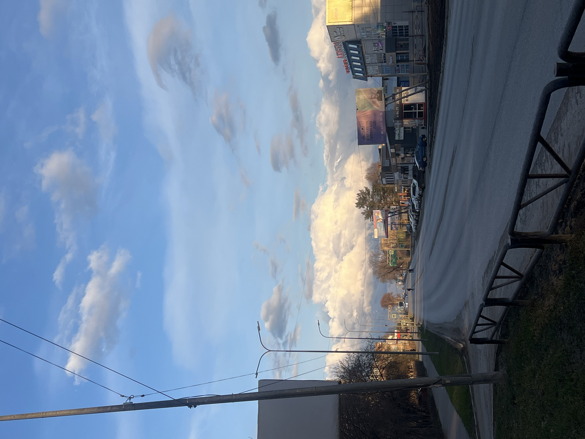 The image presents a street scene in Chaikovsky, Russia, under a dynamic, partly cloudy sky. The lighting suggests late afternoon or early evening, with long shadows cast by objects and some clouds illuminated by warm sunlight, contrasting with areas already in shadow. The sky is a clear blue, dotted with various cumulus clouds, some appearing soft and diffused, others more defined and bright.
A wide, multi-lane road, possibly wet or reflective, extends into the distance, flanked by sidewalks and a grassy verge. On the immediate right, a dark metal barrier or fence runs along the roadside. Overhead power lines and double-armed streetlights are prominent elements of the urban infrastructure. Along the left side of the street, several buildings of varying heights and designs are visible, housing businesses and residences. Bare trees and sparse vegetation suggest a cooler season, such as late autumn, winter, or early spring.
Visible text on a large billboard on the left prominently reads "МЫ С ТОБОЙ! МИР ОДИН НА ДВОИХ! ДВОРЦОВЫЙ ГОРОД АНТАЛИЯ 2025" (We are with you! The world is one for two! Palace City Antalya 2025), indicating an advertisement, likely for travel or real estate. Other signs on buildings include partial text like "OREL" and fragmented phone numbers such as "+7-984-77". Several cars are parked along the road, and a solitary pedestrian is faintly discernible in the distance. The scene captures a typical urban environment with a strong emphasis on the dramatic sky.