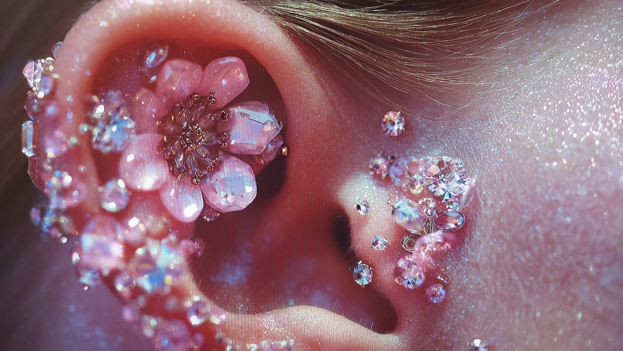A close-up, macro photograph captures an ear adorned with numerous pink, faceted crystals and glitter. A prominent, pink, floral-shaped embellishment with metallic accents is positioned within the ear canal. The skin of the ear is soft pink, with individual hairs visible near the top of the frame. The overall aesthetic is delicate and sparkling, with a bokeh effect blurring the background. The image does not depict any recognizable landmarks or features that would allow for identification of its location in Tbilisi, Georgia.