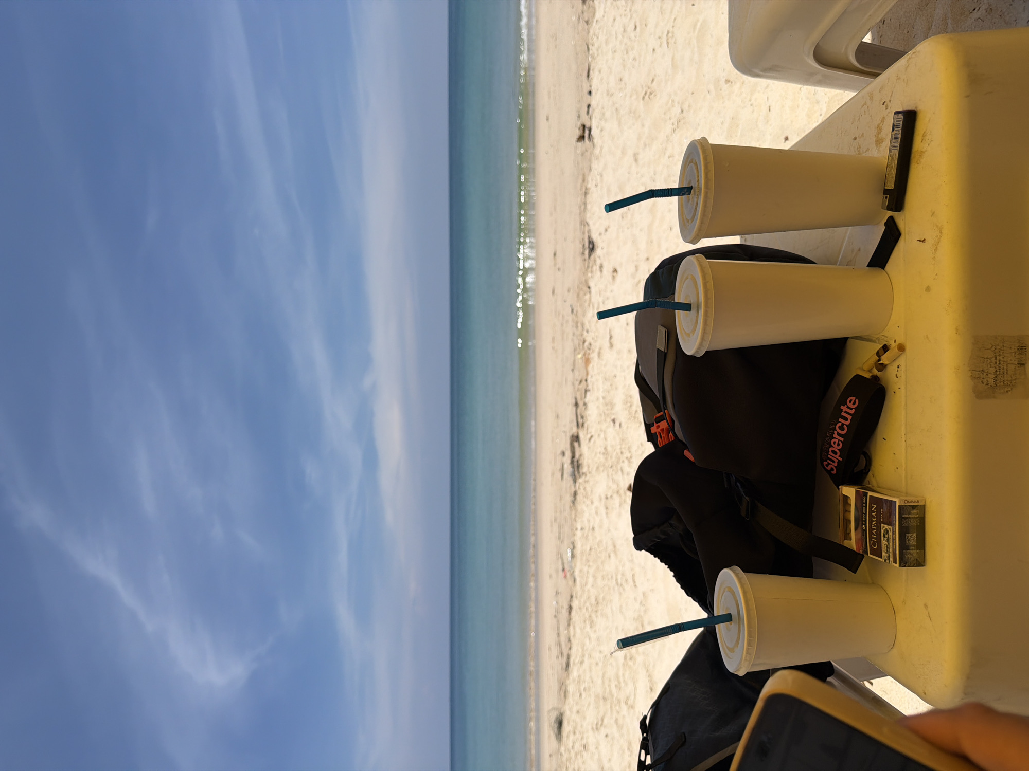 The image, rotated 90 degrees clockwise, presents a serene beach scene with the horizon running vertically down the middle. To the left of the horizon is a clear blue sky adorned with faint wispy white clouds. To the right of the horizon, the tranquil turquoise ocean meets a light sandy beach, where gentle waves can be seen along the shoreline.
In the immediate foreground, on the right side of the image, a white plastic table and parts of white plastic chairs are visible. On the table are three white disposable cups, each topped with a lid and a light blue straw, suggesting they contain refreshing beverages. A black backpack rests on the table next to the cups.
Other objects on the table include a black rectangular item, possibly a lighter or a vape, and two packs of cigarettes. The brand "CHAPMAN" is clearly visible on one of the cigarette packs. A black strap, likely belonging to the backpack, features the text "Supercute." At the very bottom right, a human hand is visible, holding a smartphone.
The scene depicts a bright, sunny daytime setting, suggesting a relaxed atmosphere. The calm waters, clear sky, and sandy environment evoke a sense of leisure and peace, indicative of a beach vacation in Choeng Thale, Thailand.