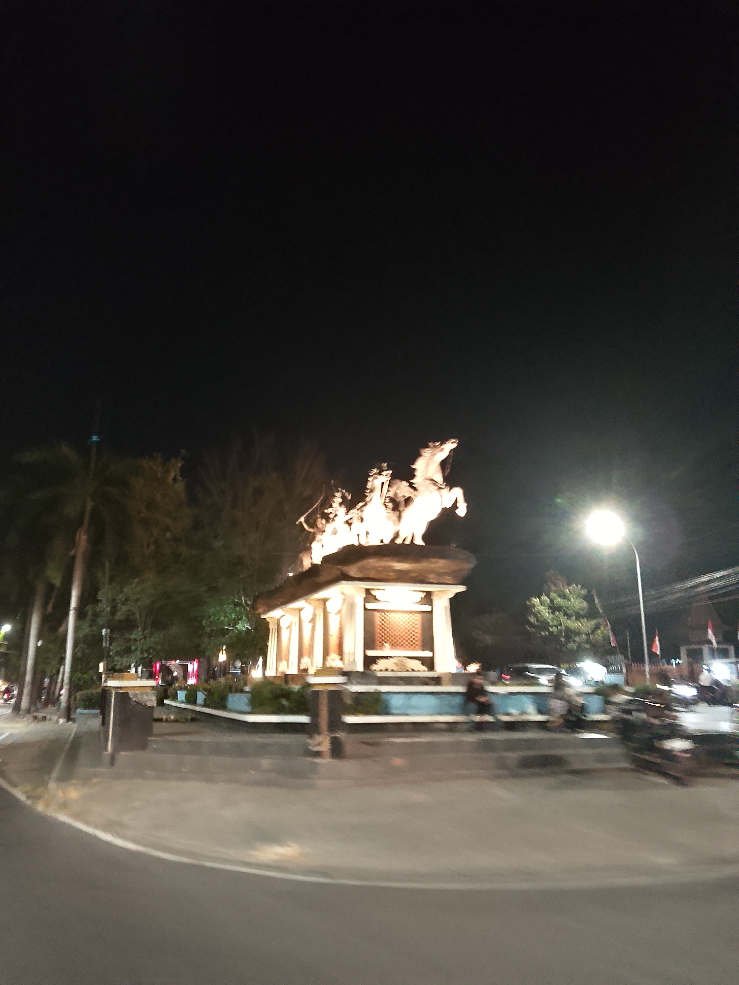 The image shows a nighttime view of a monument in Balun, Indonesia. The monument, illuminated by warm light, features a sculpture of several figures riding horses on top of a pedestal. The pedestal is adorned with pillars and has a textured grate-like design on its front. Surrounding the monument is a paved area with a road curving in the foreground. Trees and streetlights are visible in the background. There are blurred figures of people and vehicles in the scene, suggesting a public space or street.