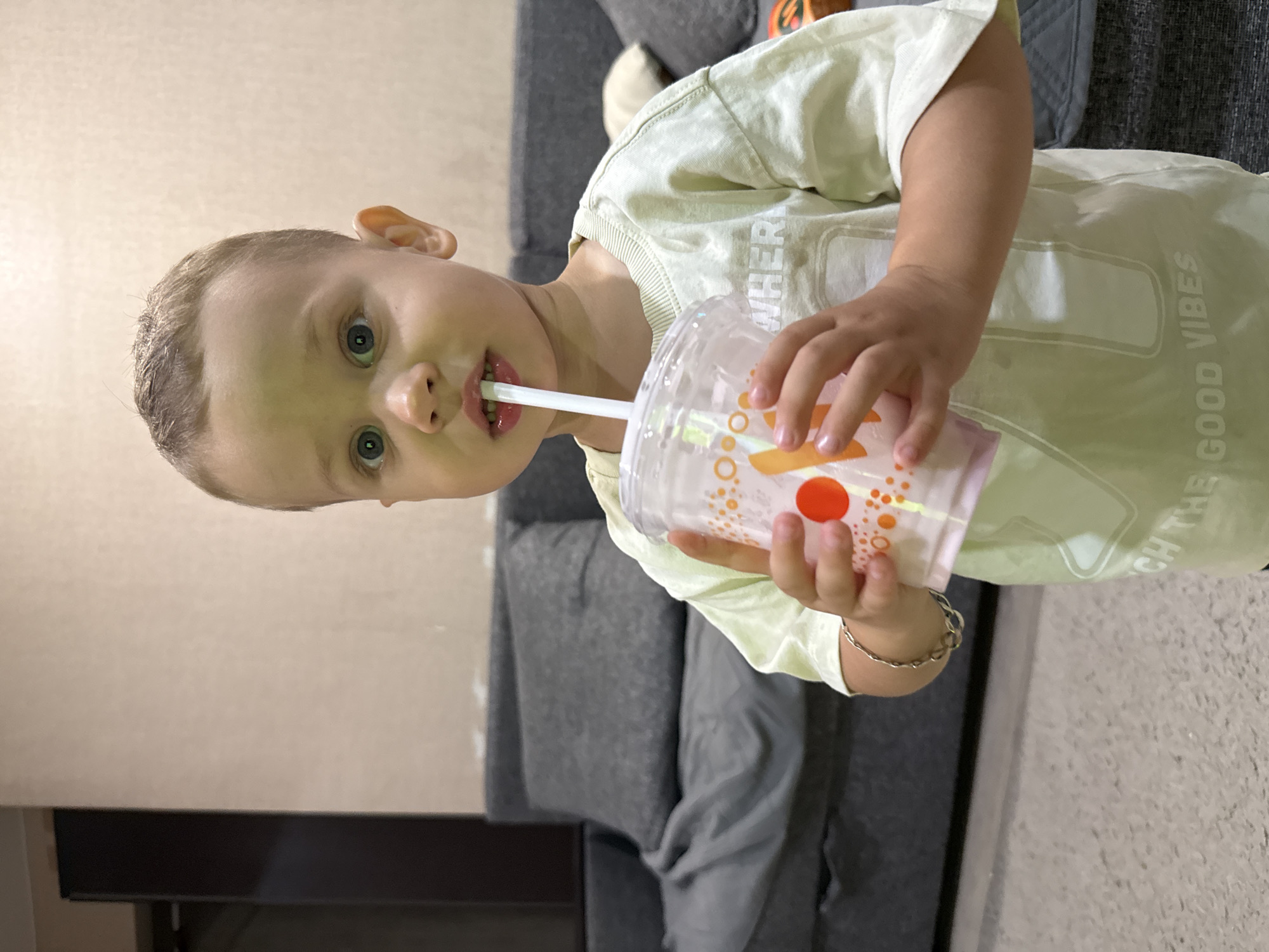 A young boy with blond hair and blue eyes is drinking from a clear plastic cup with a white straw. He is wearing a light green t-shirt with the words "WHERE" and "GOOD VIBES" printed on it. The boy is standing in a room with a gray sofa and a beige wall. The image appears to have been taken in a residential setting, possibly in the city of Rusanovo, Russia.