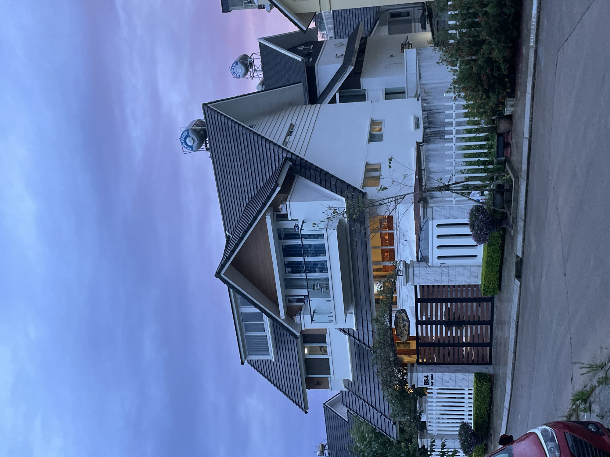 The image depicts a modern villa in Da Lat, Vietnam, at dusk. The villa is a multi-story structure with a distinctive dark-tiled, pitched roof. The exterior walls are primarily white with some wooden accents. Two large, blue water tanks are visible on the roof. The villa is surrounded by a white picket fence and a decorative gate made of wood and stone. Lush green bushes and potted plants adorn the front yard. In the foreground, a red car is partially visible on the street. The sky above is a gradient of purple and blue, indicating twilight. The overall scene conveys a sense of serene, upscale residential living. The sign at the entrance reads "B54 Golf Valley".