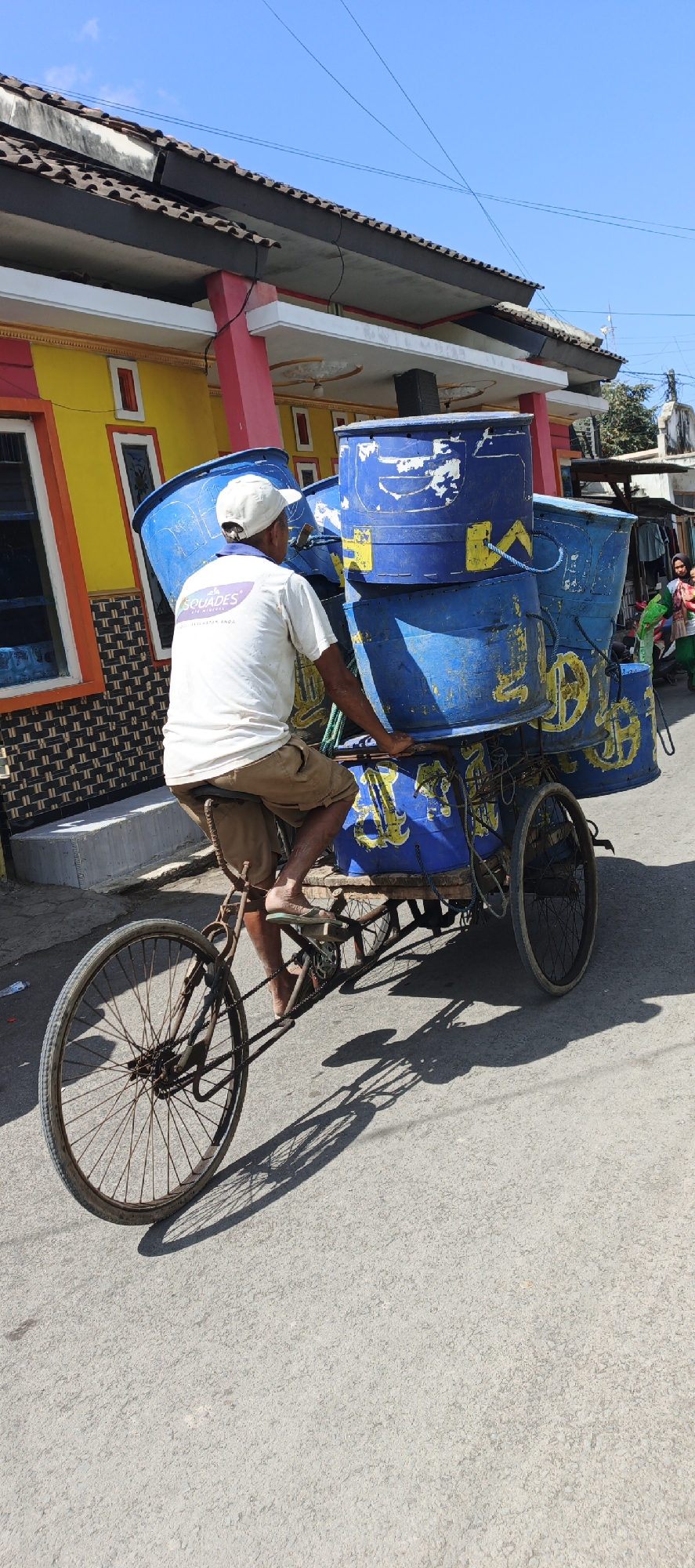 A man wearing a white cap, white t-shirt with a logo, and brown shorts rides a cargo bicycle laden with several large blue barrels stacked on a wooden platform. The bicycle has a single front wheel and two rear wheels. The man's feet are bare, and he is wearing sandals. The barrels are blue with yellow markings and appear to be made of plastic. The background shows a yellow and red building with a tiled roof. The scene is set on a paved street under a clear blue sky. The image was taken in Puger, Indonesia, as indicated by the location tag. The sunlight casts strong shadows from the bicycle and barrels onto the street. There are other people visible in the background, including a woman in a green headscarf and a child.