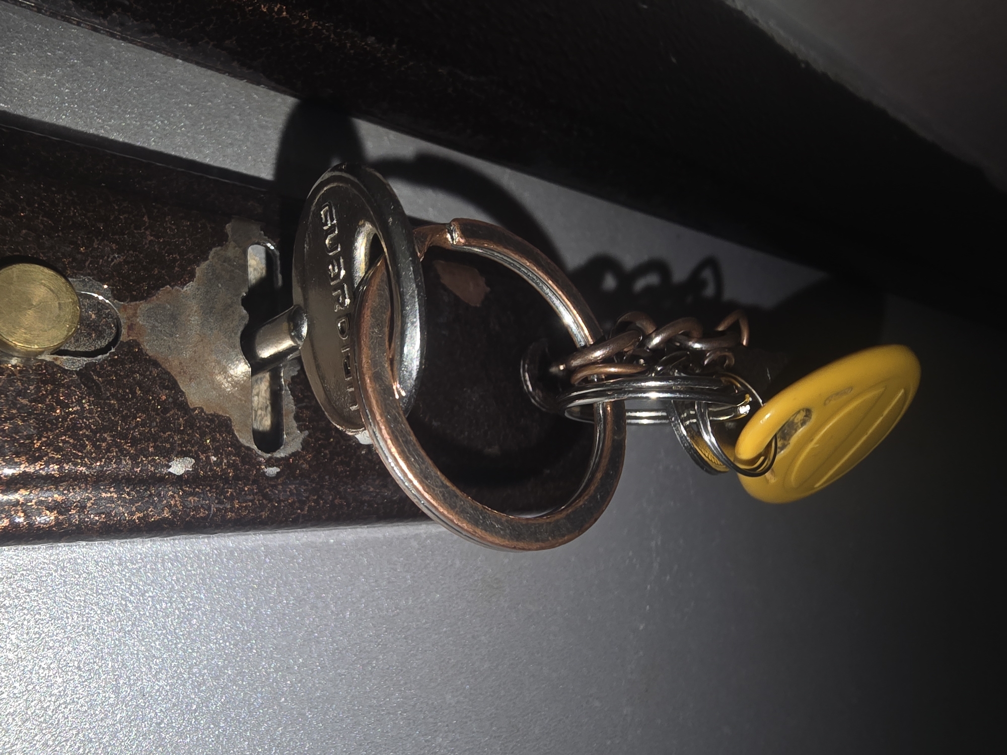 The image shows a close-up of a metal door lock mechanism with a set of keys hanging from it. The lock appears to be on a dark, possibly metal, door with a textured, reddish-brown surface. A key with the word "GUARDIAN" visible on its head is attached to a large, circular keyring. Several smaller keys and a yellow tag are also attached to the keyring via a chain. The background is a mix of light gray and dark, out-of-focus areas. There are no people or animals visible in the image. The image is a still photograph with no discernible action or interaction occurring. The scene is static, focused on the detail of the lock and keys. There is no information in the image that would allow for a determination of its location within the city of Ulan-Ude, Russia.
