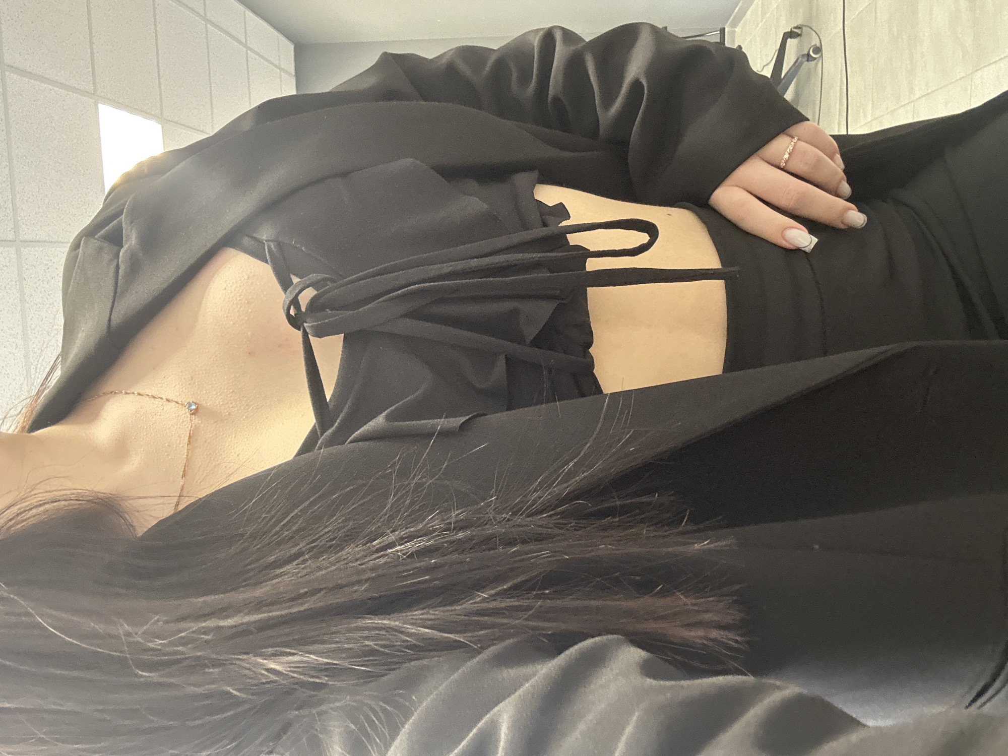 An indoor, waist-up photograph depicts an individual, likely female, standing in what appears to be a commercial or office building. The precise location within Izhevsk, Russia, cannot be determined from the visual information provided.
The individual wears a black open blazer over a black crop top with prominent tie-front or drawstring detailing, exposing a portion of the midriff. Black bottoms, possibly a skirt or trousers, are visible from the waist down. Long, dark hair falls over the individual's left shoulder and chest. A delicate gold-colored necklace with a small, light blue, heart-shaped pendant is worn around the neck. The right hand, with manicured nails featuring light polish and a thin dark line design, rests on the hip/lower abdomen. A gold-colored ring with small stones is on the ring finger of the right hand.
The background shows a white suspended ceiling with visible acoustic tiles and integrated rectangular fluorescent light panels, one of which is illuminated. The walls are painted a light grey or blue. The floor is covered with light-colored square tiles, suggesting a grid pattern. A dark object, possibly a chair back or furniture, is partially visible in the far right background. The scene is brightly illuminated by artificial lighting. No other individuals or interactions are visible.