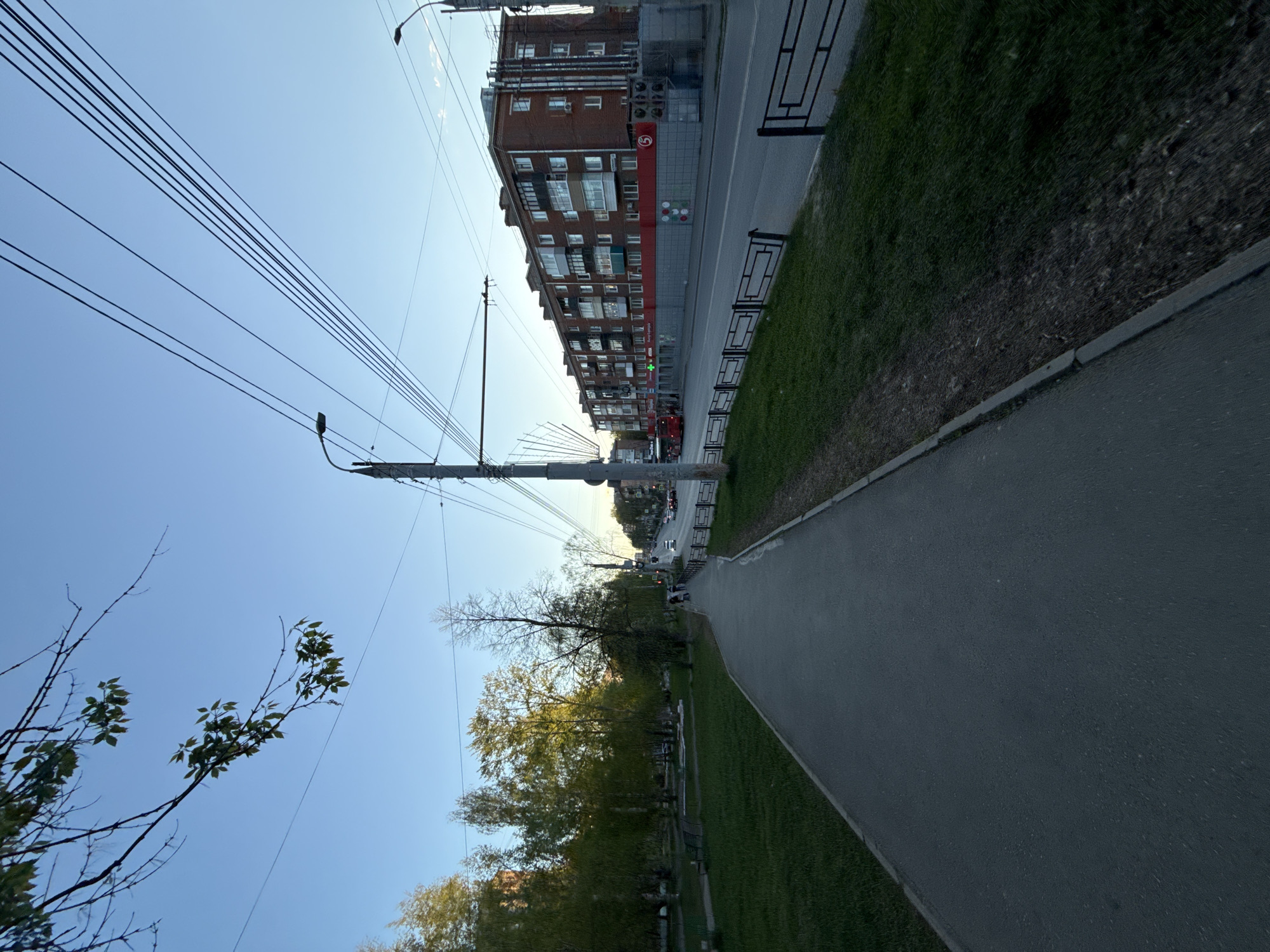 The image shows a sidewalk in a city, likely Izhevsk, Russia, based on architectural style. The sidewalk is paved and runs alongside a grassy area with trees. A building stands to the right of the sidewalk, featuring a red and white storefront with a Cyrillic script. Power lines stretch across the sky above the street, and a few cars are visible further down the street. There is no visible interaction between any people.