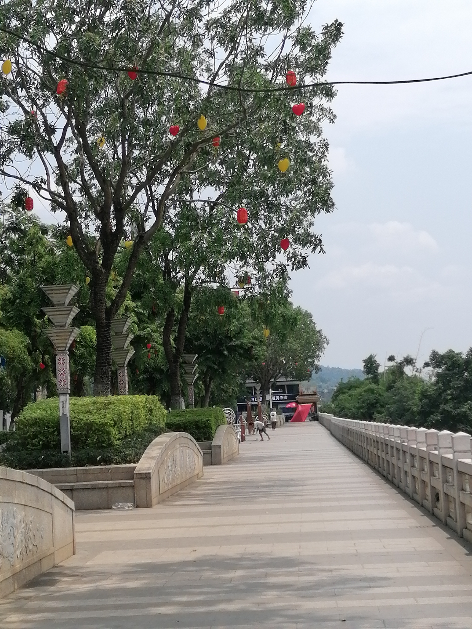 A pedestrian walkway stretches into the distance, flanked by a decorative stone railing on the right. On the left, a tree adorned with red and yellow lanterns overlooks a landscaped area with trimmed bushes and ornate lampposts. Two people are present on the walkway, appearing to be involved in cleaning or maintenance activities. The walkway leads towards a building with a red awning. In the background, a hillside with trees is visible beneath a partly cloudy sky. The location is identified as Hekou, China.