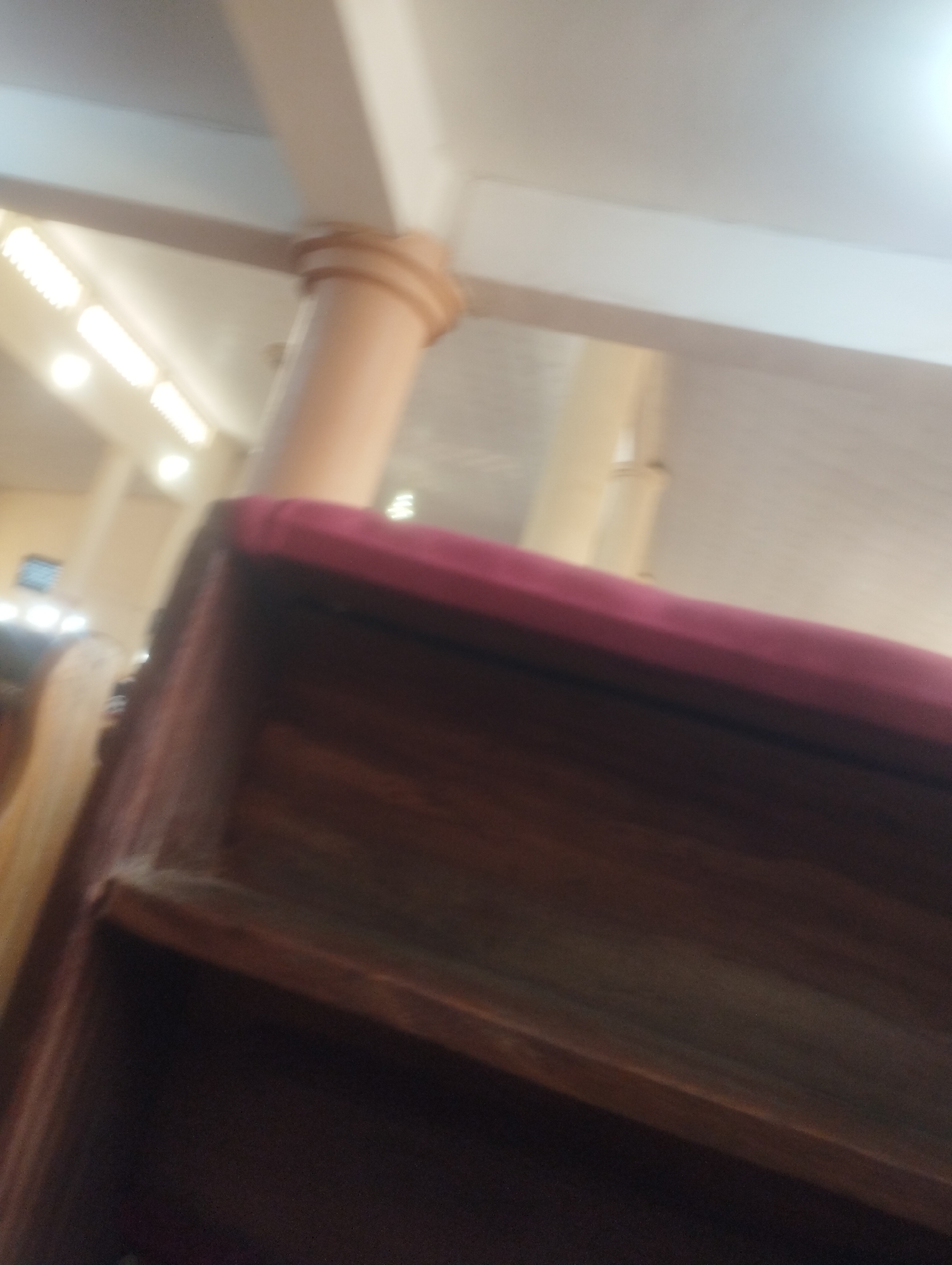 The image shows an interior view of a building, possibly a church or a community hall, in Jos, Nigeria. The scene is dimly lit, with fluorescent lights visible along the ceiling. In the foreground, a dark wooden bench with a maroon cushion is partially visible. The bench appears to be part of a seating arrangement, with other wooden structures and seating visible in the background. A large, beige-colored pillar is also prominent in the middle ground, supporting the ceiling structure. The overall impression is of an indoor public space, possibly with a religious or communal purpose.